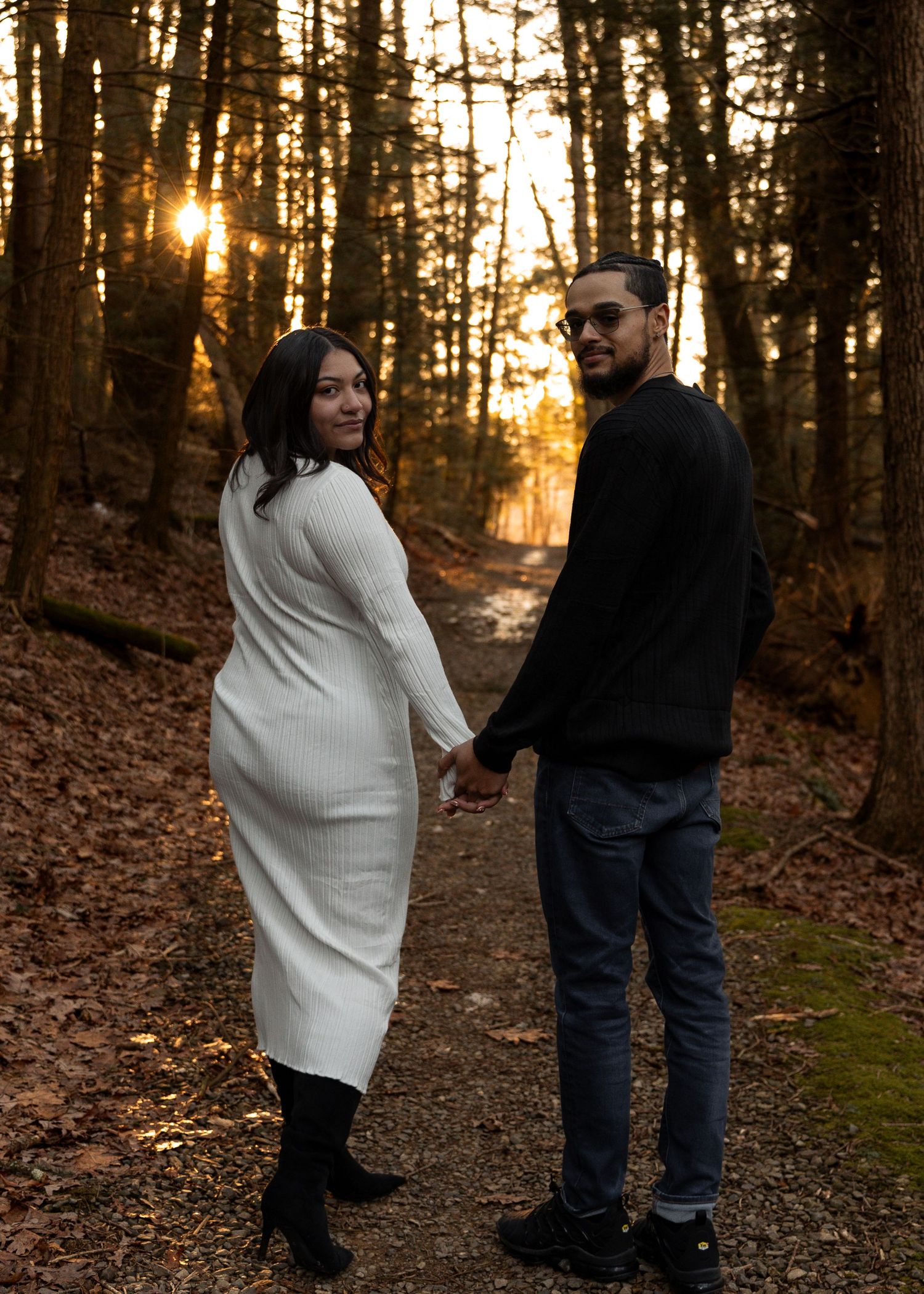 Couple holding hands on forest path at golden sunset, woman in white knit dress and man in black outfit.