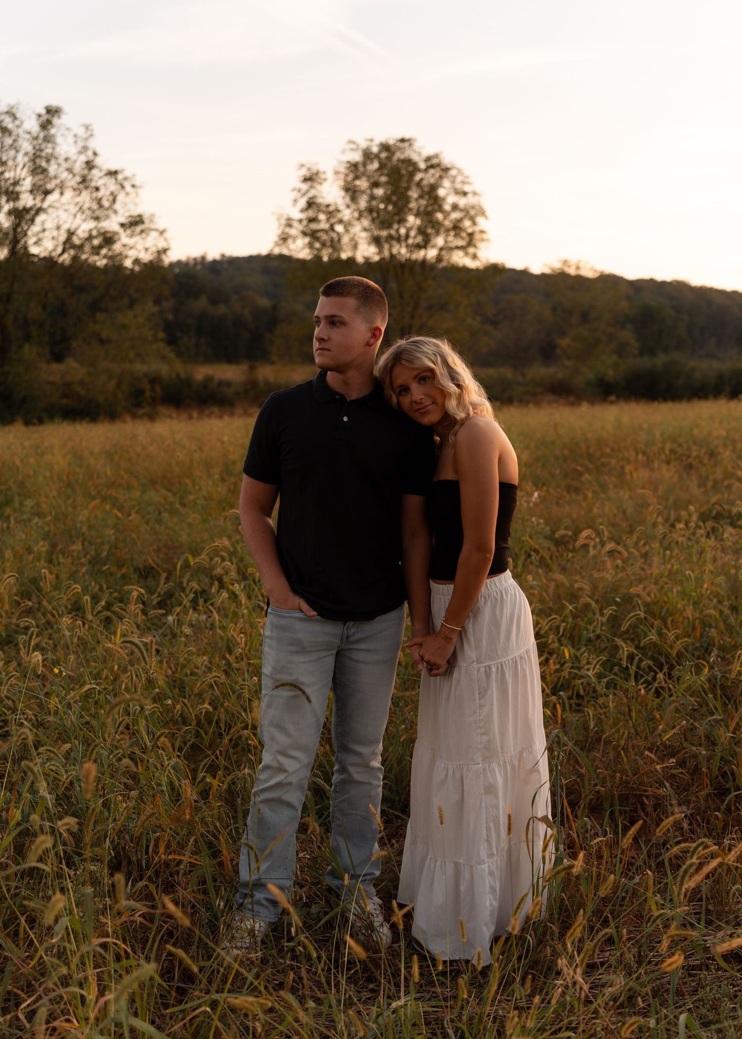 Romantic couple holds hands in a golden field at sunset, woman in white skirt leans on mans shoulder.