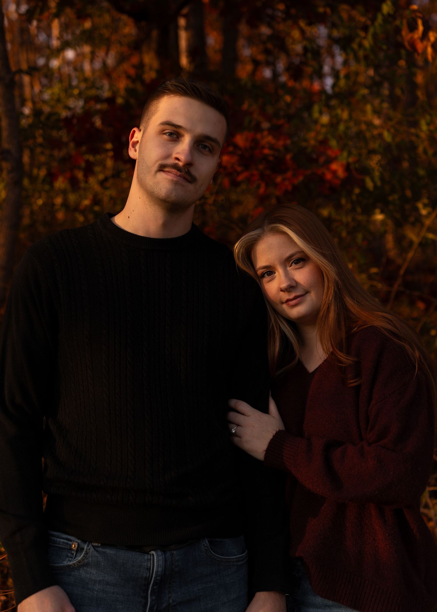 Engaged couple poses together among autumn foliage at dusk, woman in burgundy sweater shows ring.