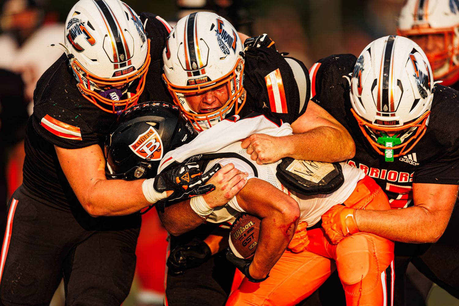 Football players in orange and white uniforms tackling an opponent during an intense game under bright stadium lights.
