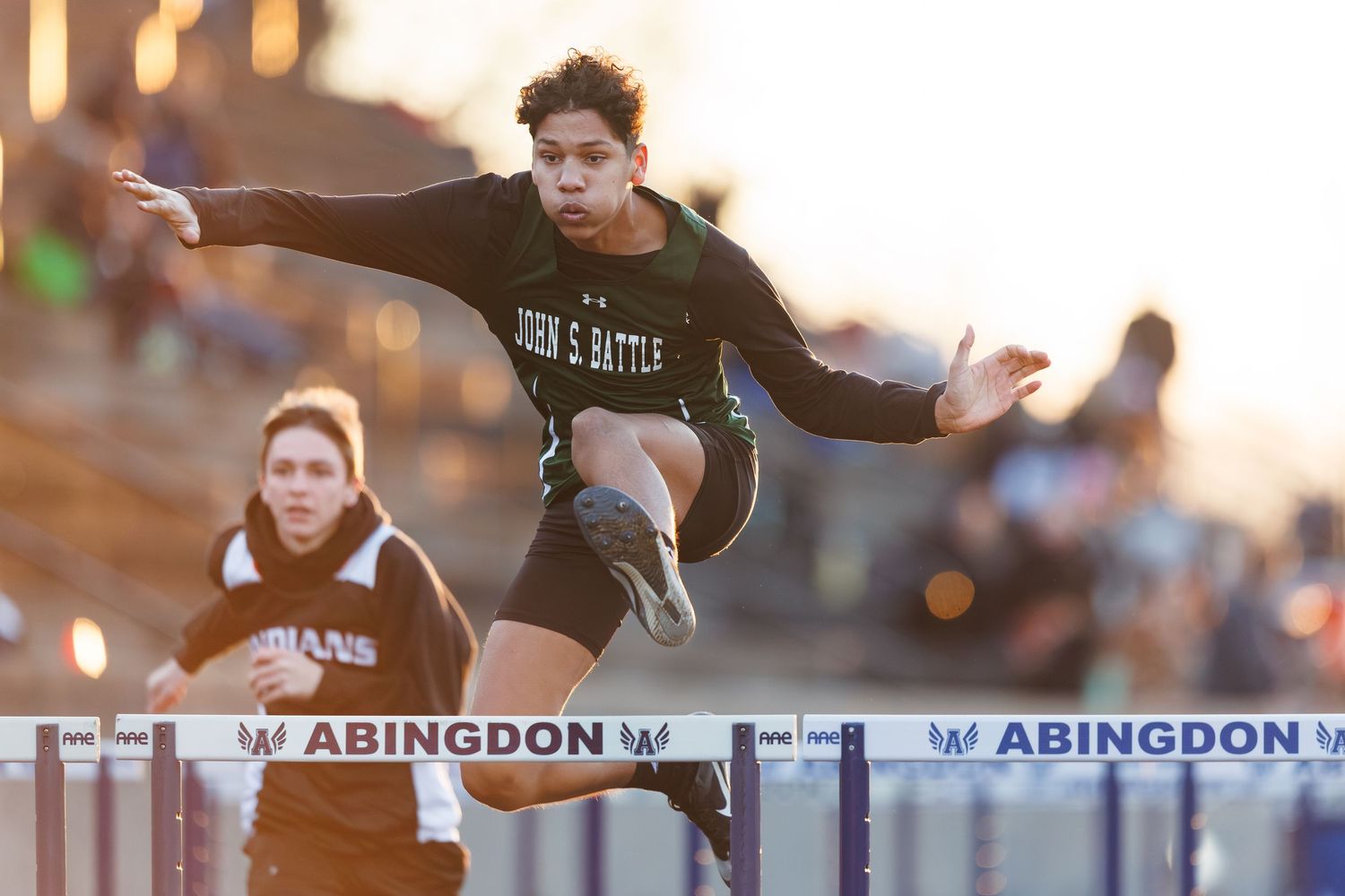 Track athletes clearing hurdles during competitive race at sunset.
