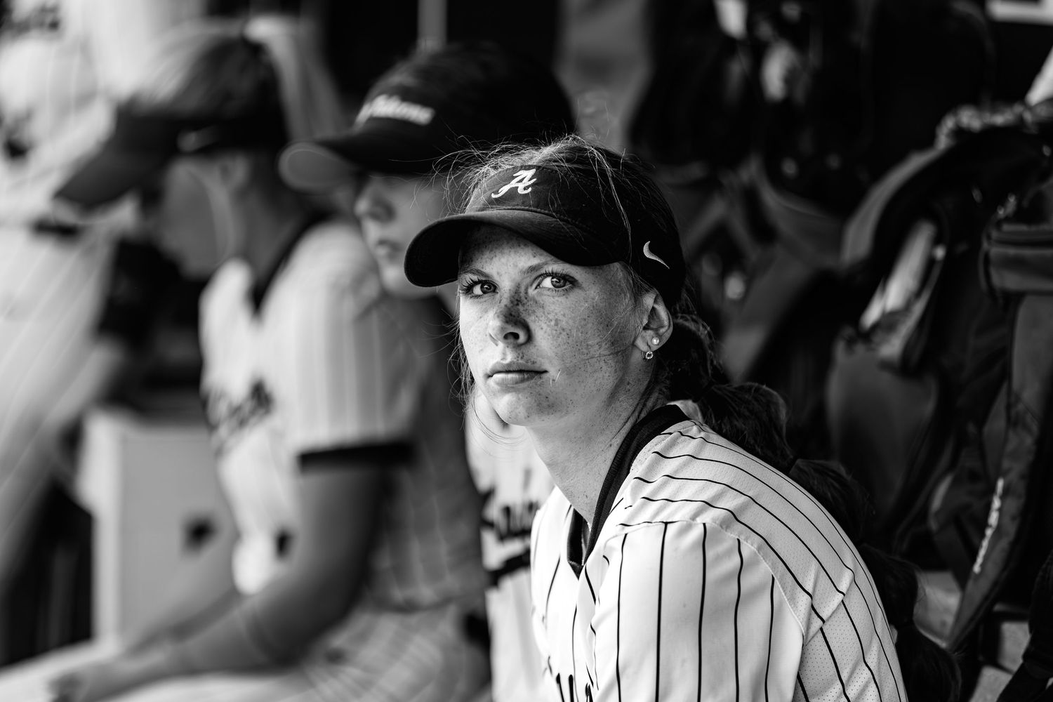 Black and white photo of a focused female baseball player in dugout wearing pinstripe jersey and cap.