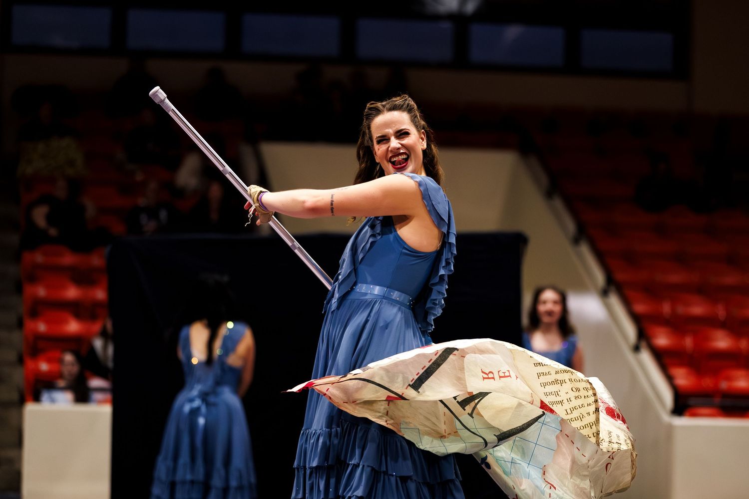 Woman in blue gown performs with a pole and prop during an indoor color guard or theatrical performance.