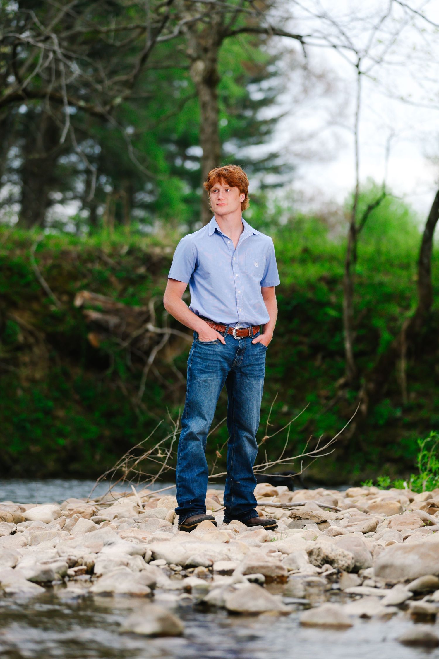Portrait of a person standing on rocks by a stream in a wooded setting.