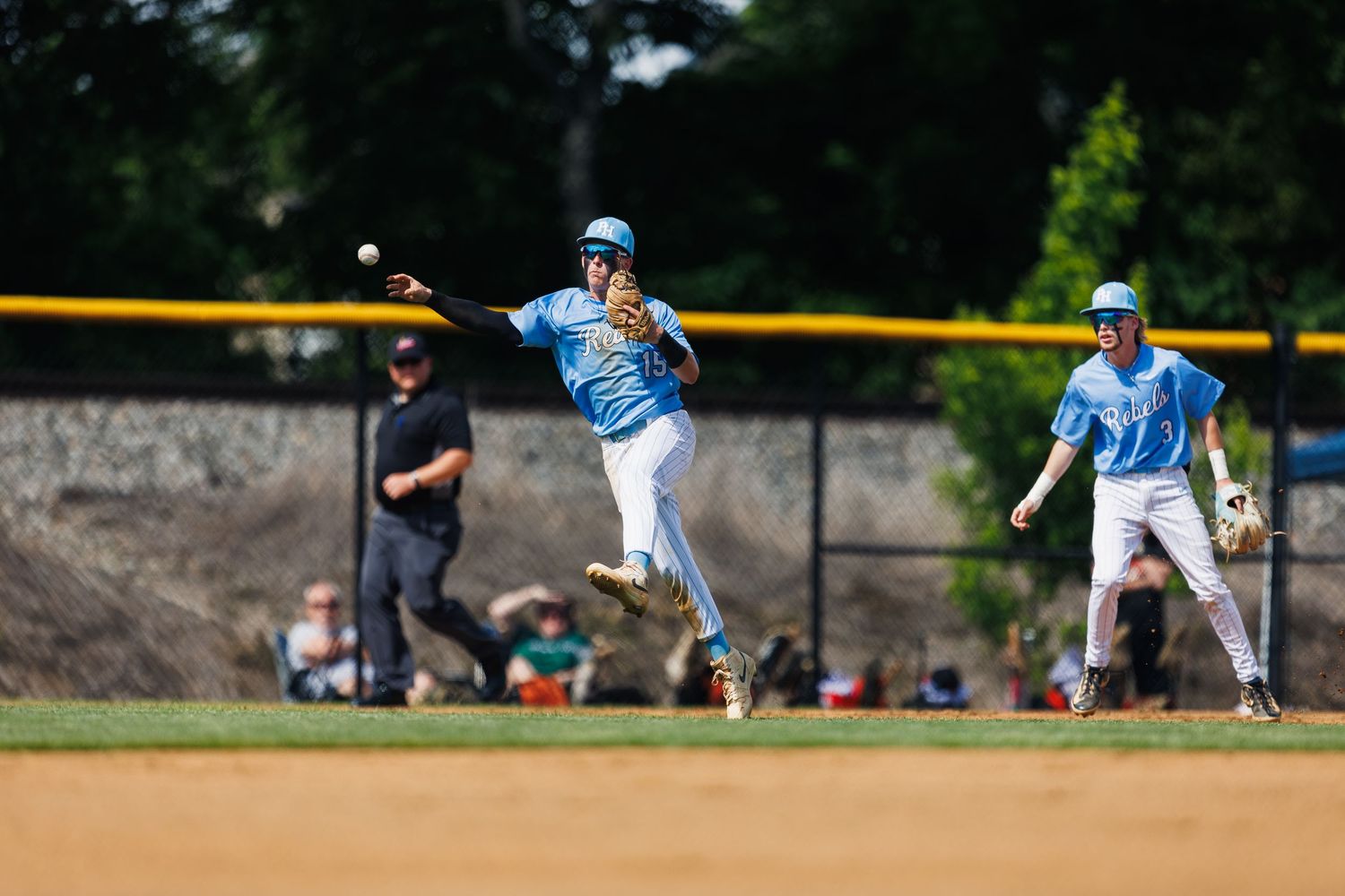 Baseball player in blue uniform leaping to throw ball during a sunny outdoor game, with teammate and umpire in background.