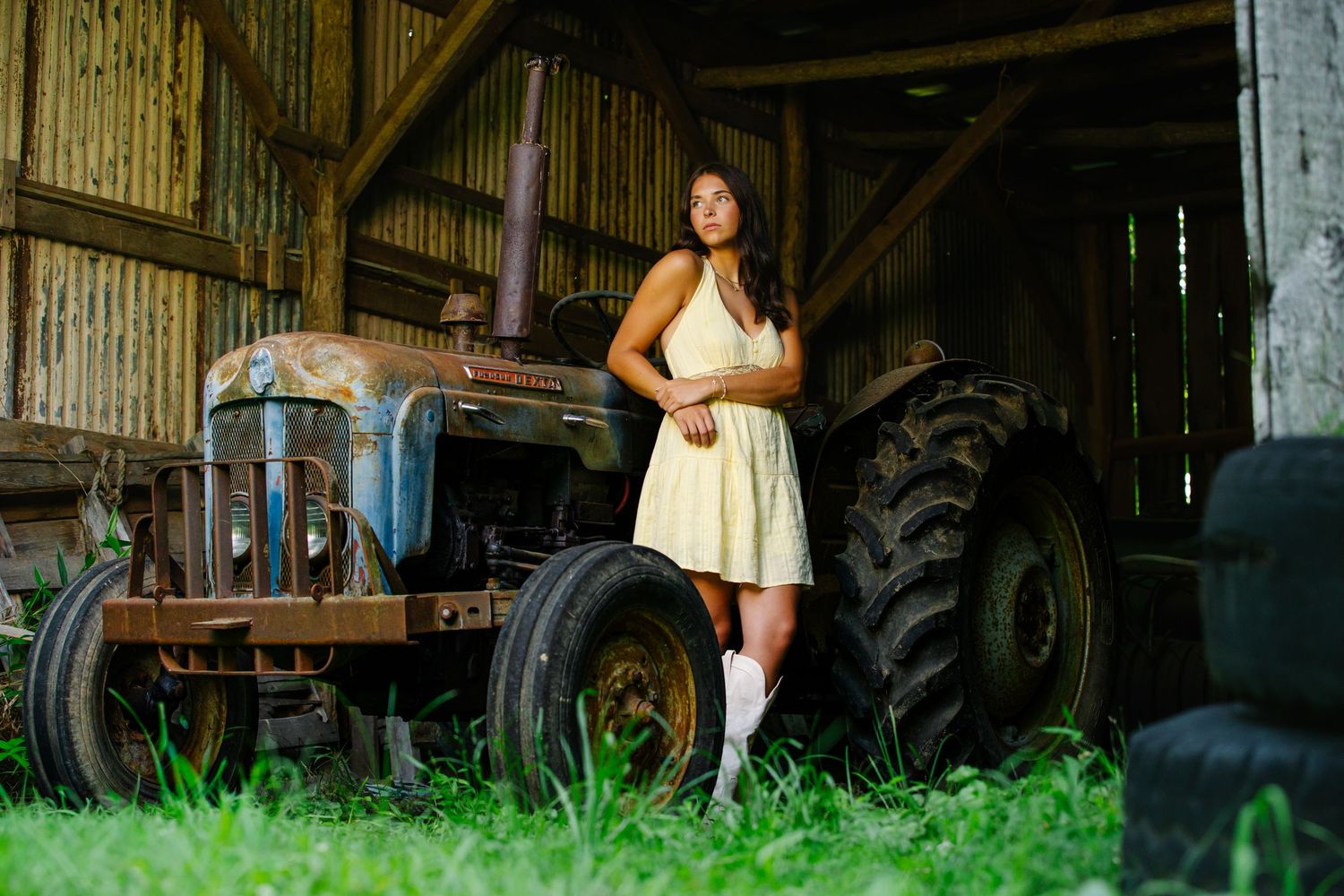 Woman in white dress and boots posing beside vintage rusty tractor inside weathered barn with green grass.