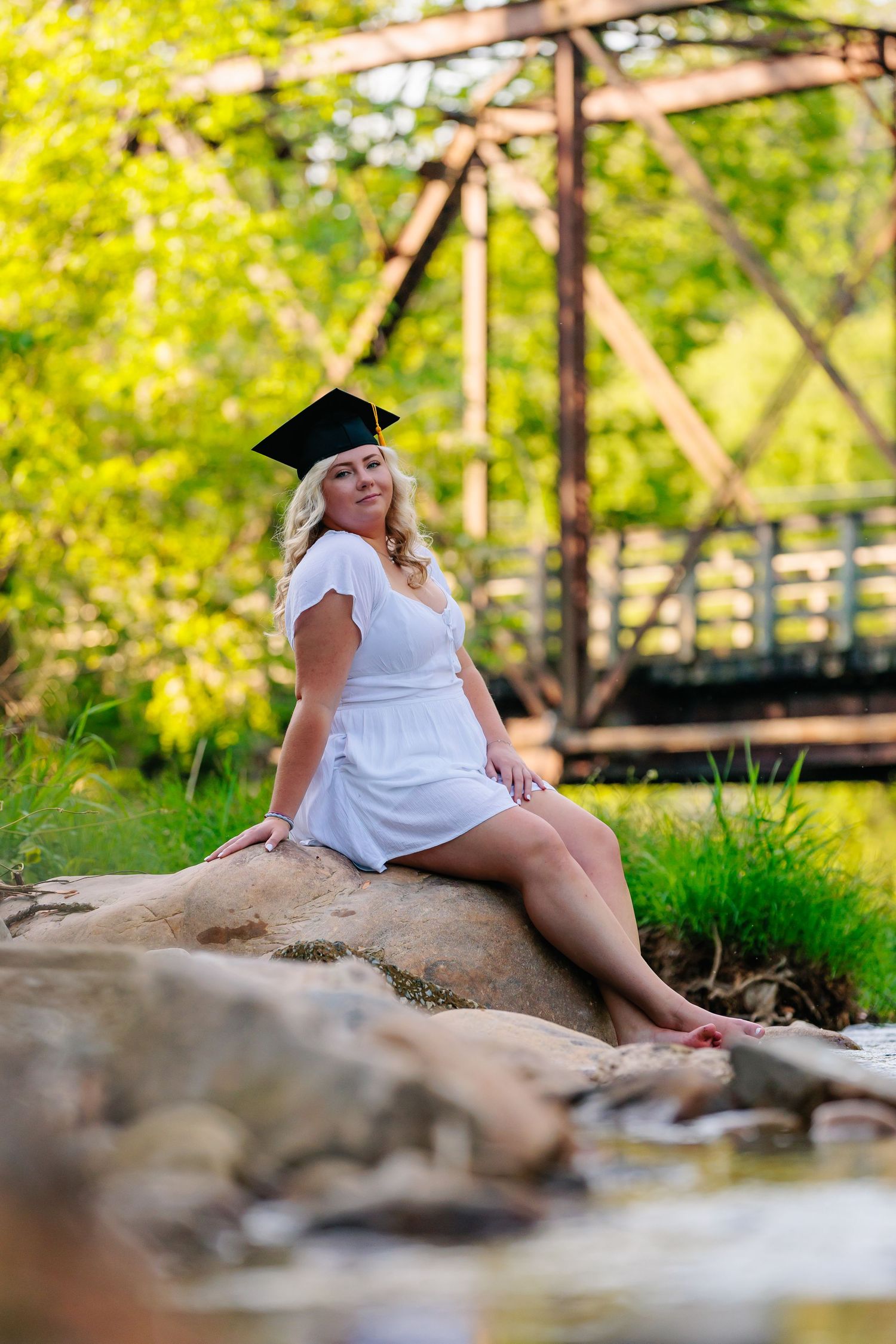 Graduate in white dress poses by creek with wooden bridge in background.