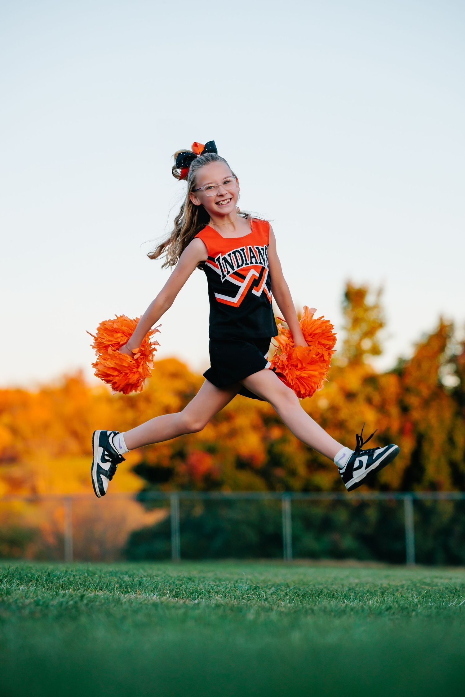 Cheerleader in orange and black uniform performs dynamic jump against autumn backdrop.