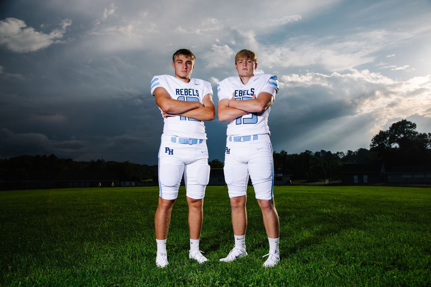 Two male football players in white Rebels uniforms posing on field under dramatic cloudy sky.
