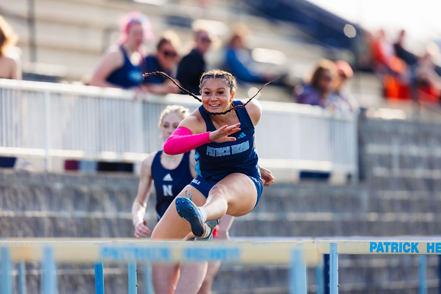 Female athlete clears a hurdle during a track and field competition at Patrick Henry stadium.