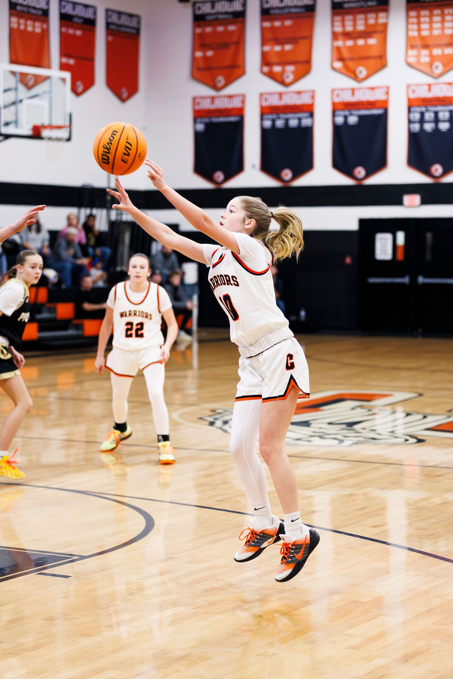 Basketball players leap for the ball during an indoor game with championship banners visible.