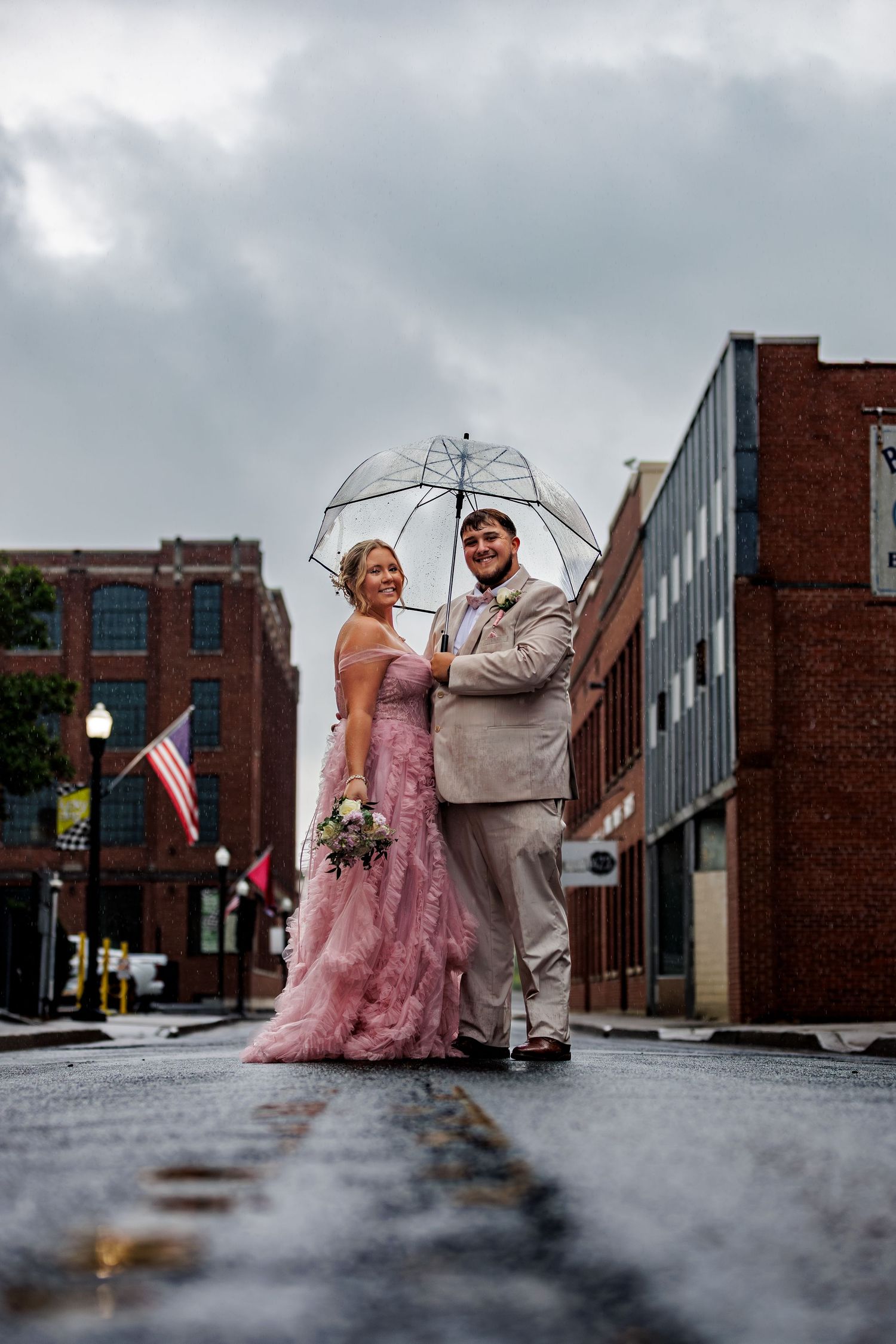 Couple in pink gown and tan suit sharing clear umbrella on rainy downtown street during wedding photo session.