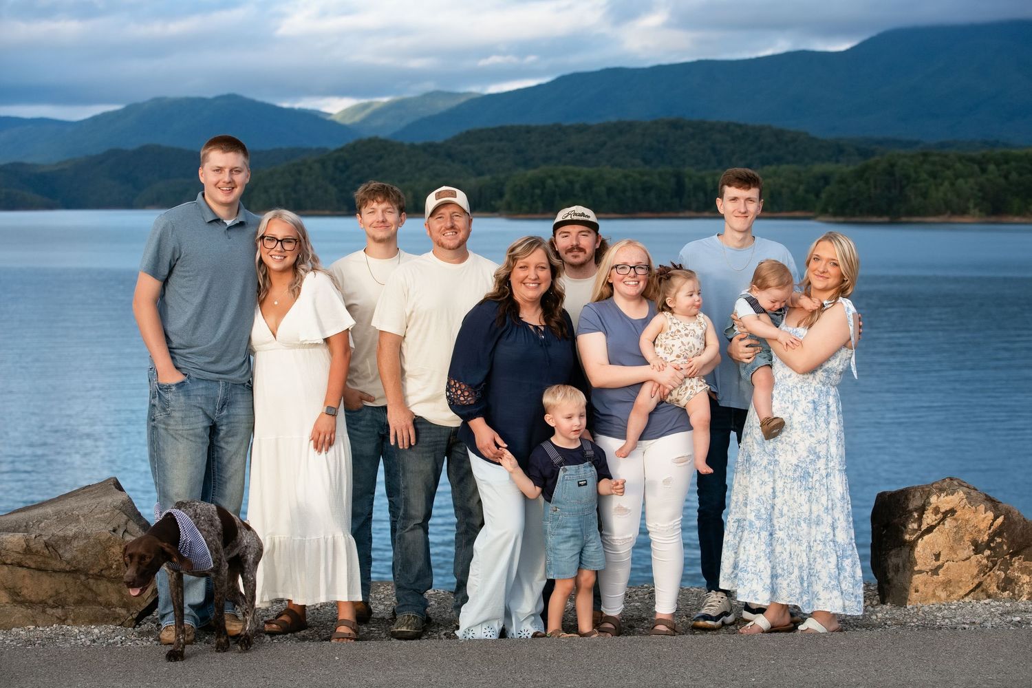 Large family group posing together near a scenic mountain lake with blue water and misty peaks in background.