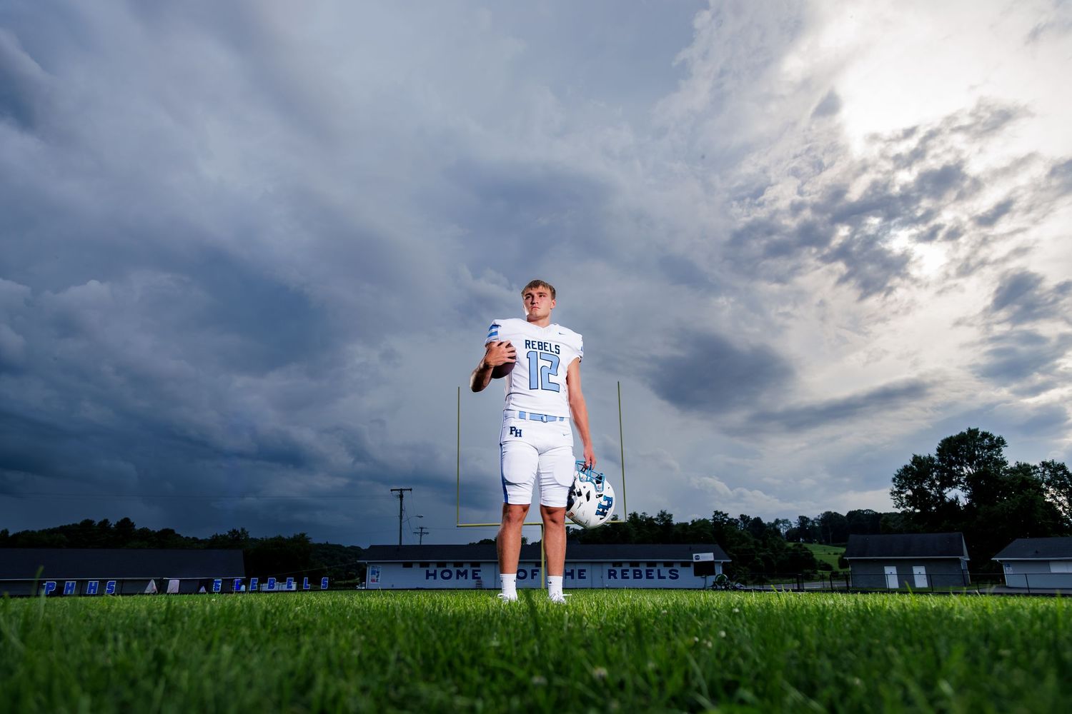 A young male soccer player stands on a green field holding a ball under dramatic stormy skies.