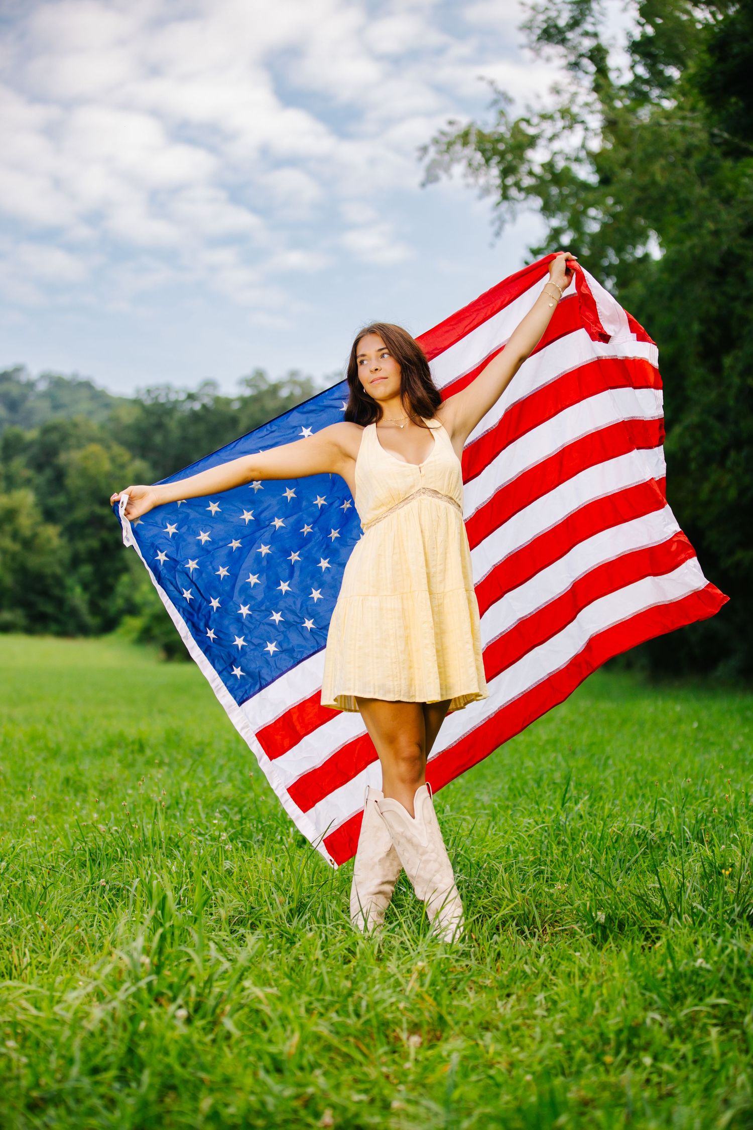 Young woman in yellow dress and cowboy boots holding an American flag in a green field on a sunny day.