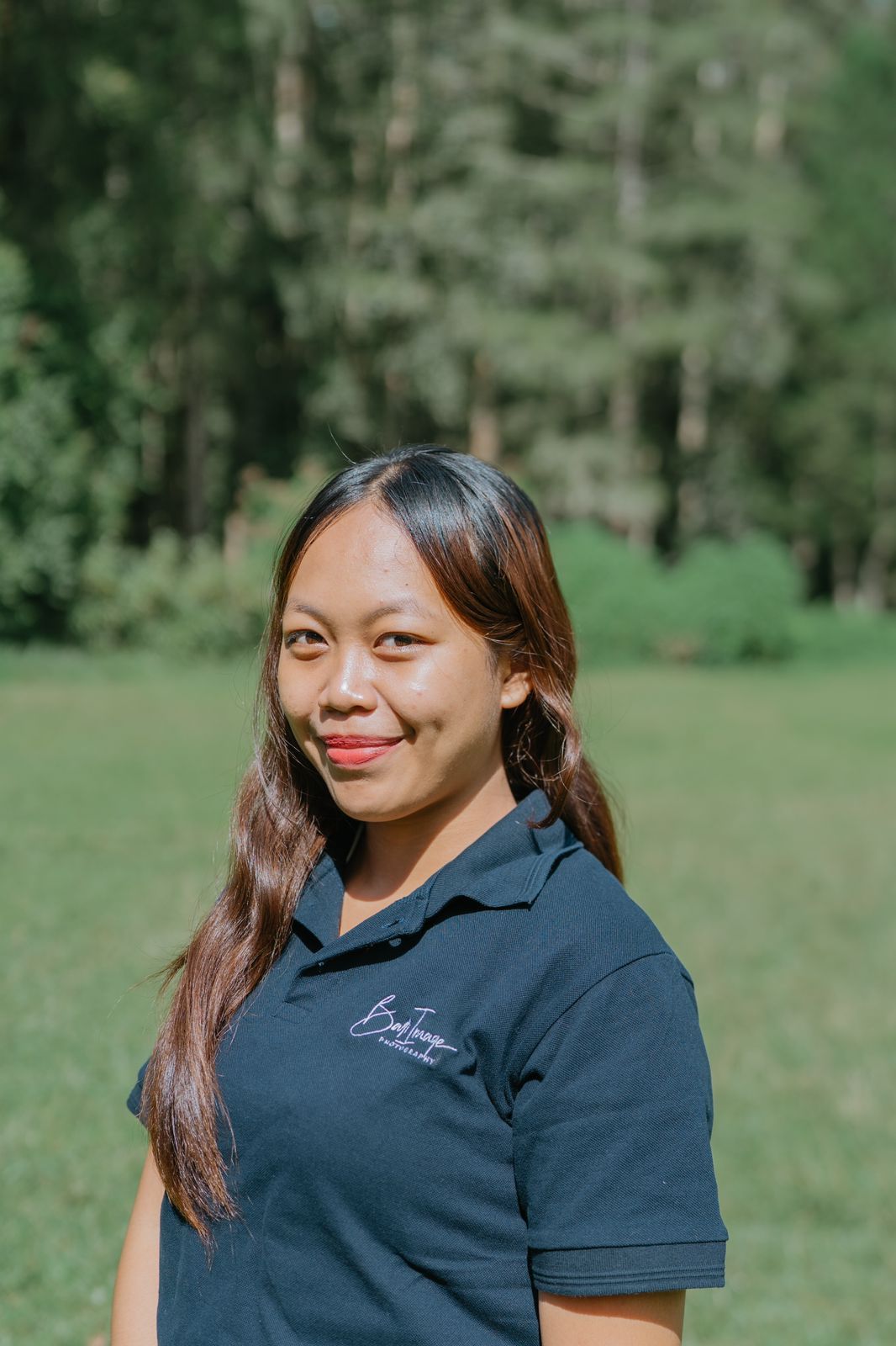 A person wearing a black polo shirt smiles warmly while standing outdoors against a green forest background.