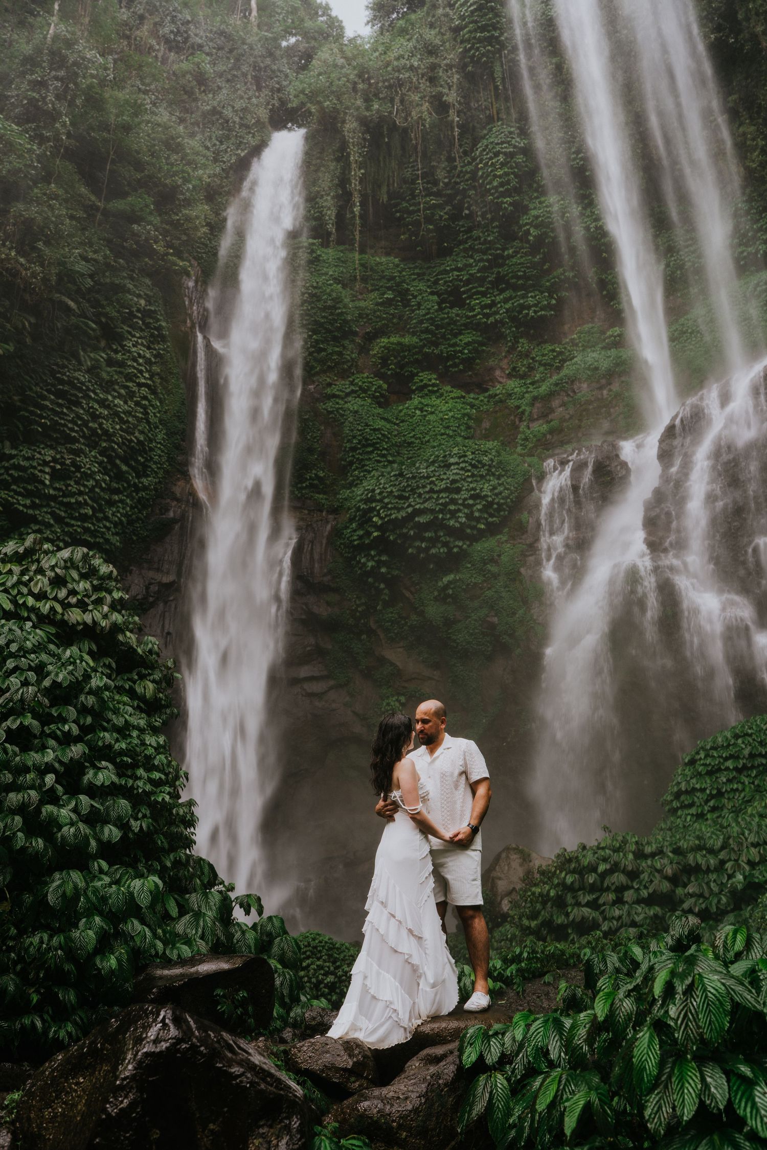 Couple posing in front of a majestic double waterfall in a tropical forest setting.
