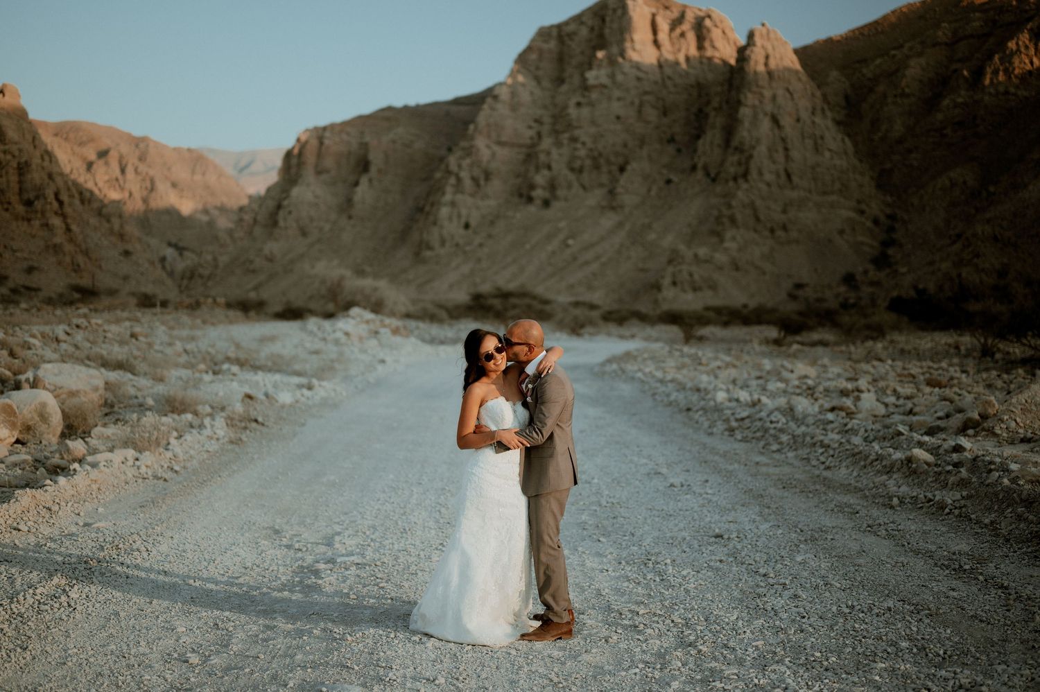 Newlyweds share romantic moment on desert road with towering cliffs behind them.