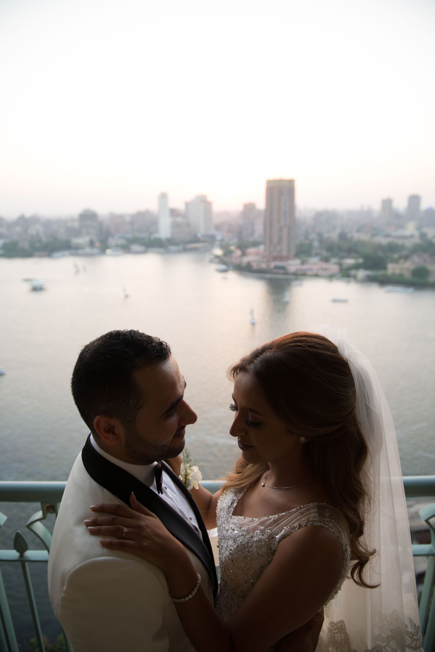 A romantic wedding couple shares an intimate moment on a balcony overlooking a city skyline at sunset.