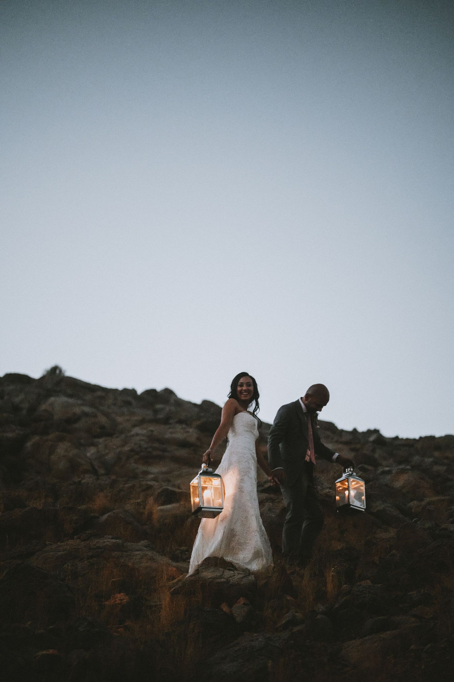 Silhouetted figures hold lanterns while standing on a rocky hillside at twilight.