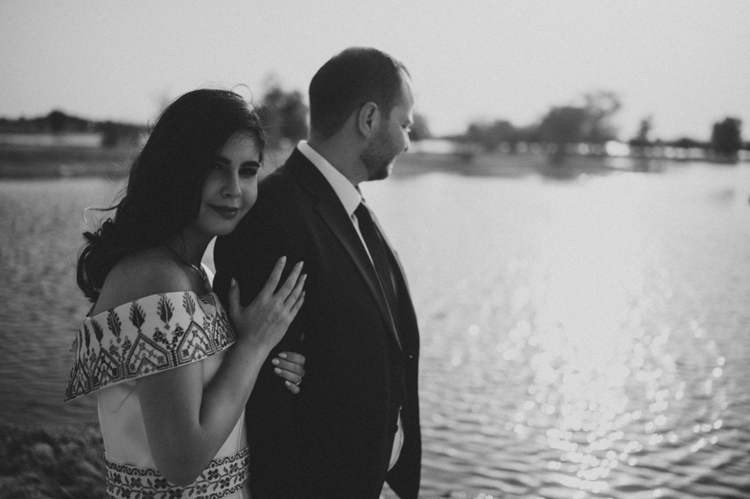 Couple in formal attire embracing by waterfront during golden hour.