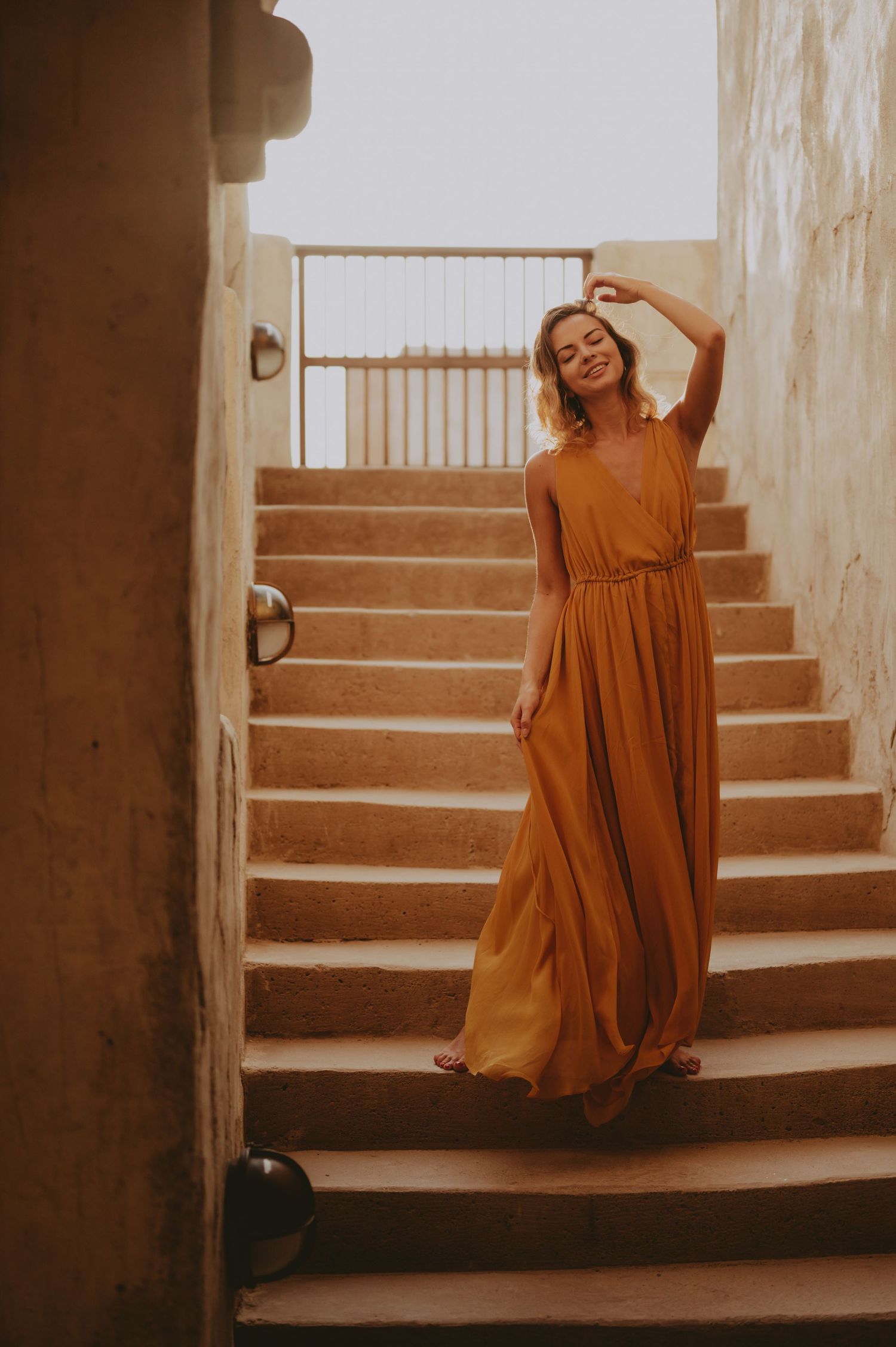 A flowing ochre dress creates dramatic movement on stone steps in warm, moody lighting.