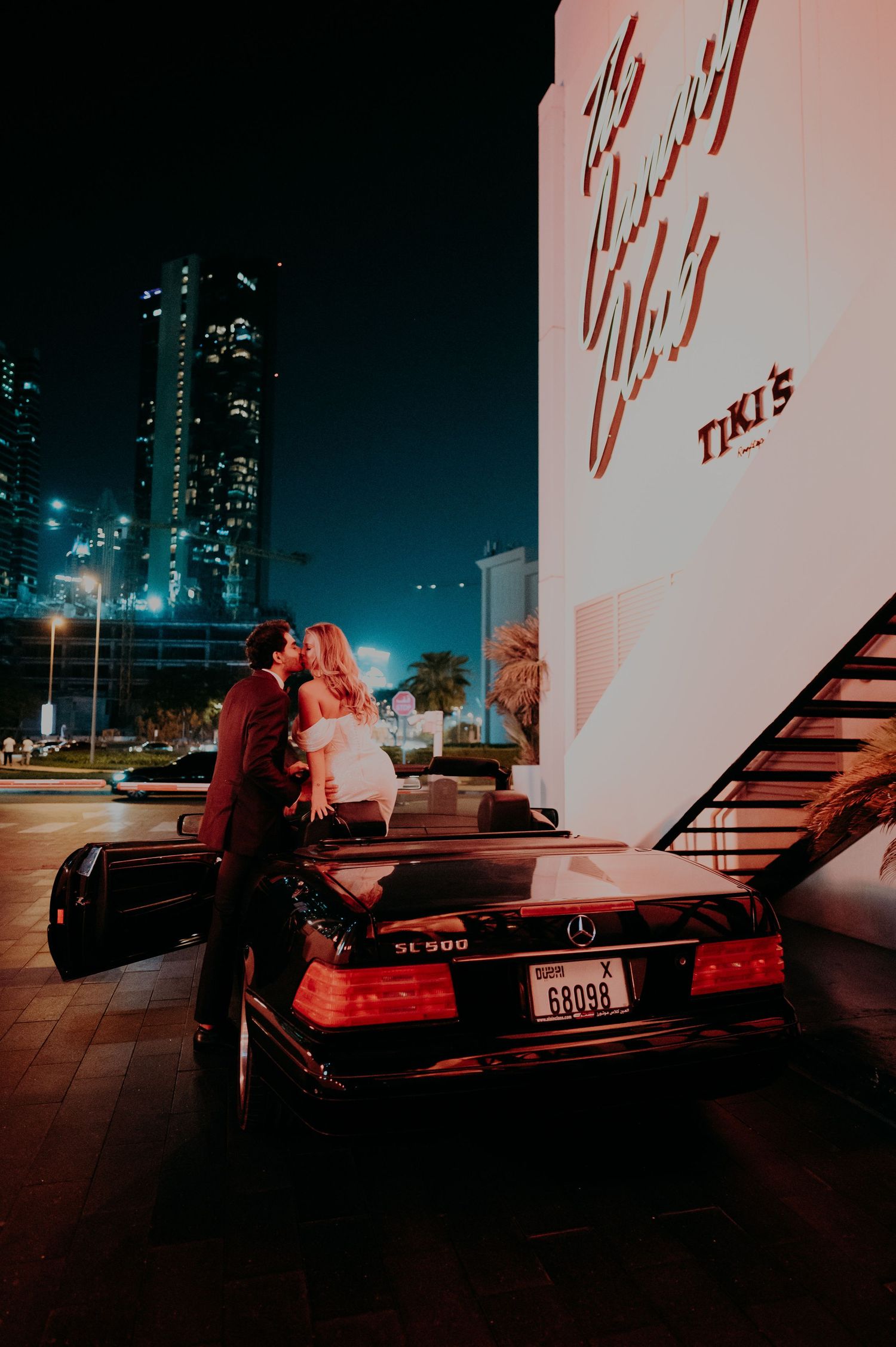 Nighttime wedding photo with couple by vintage car under neon lights.