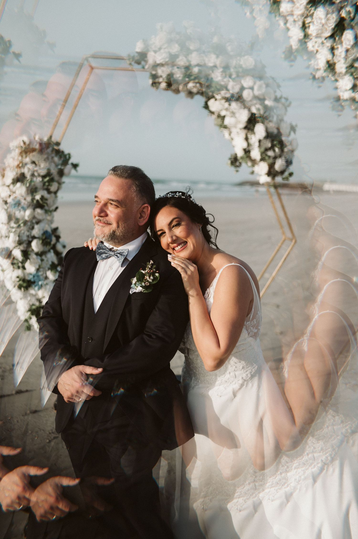 A romantic wedding couple shares joyful laughter under a floral arch with white blossoms.