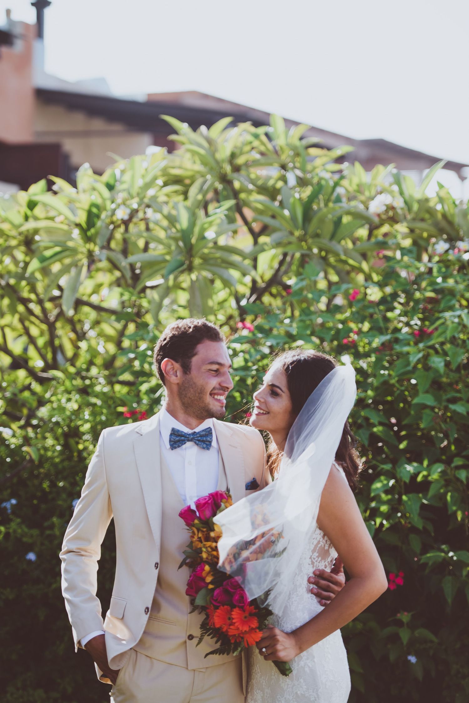 A couple in wedding attire embraces outdoors against a backdrop of lush green foliage.