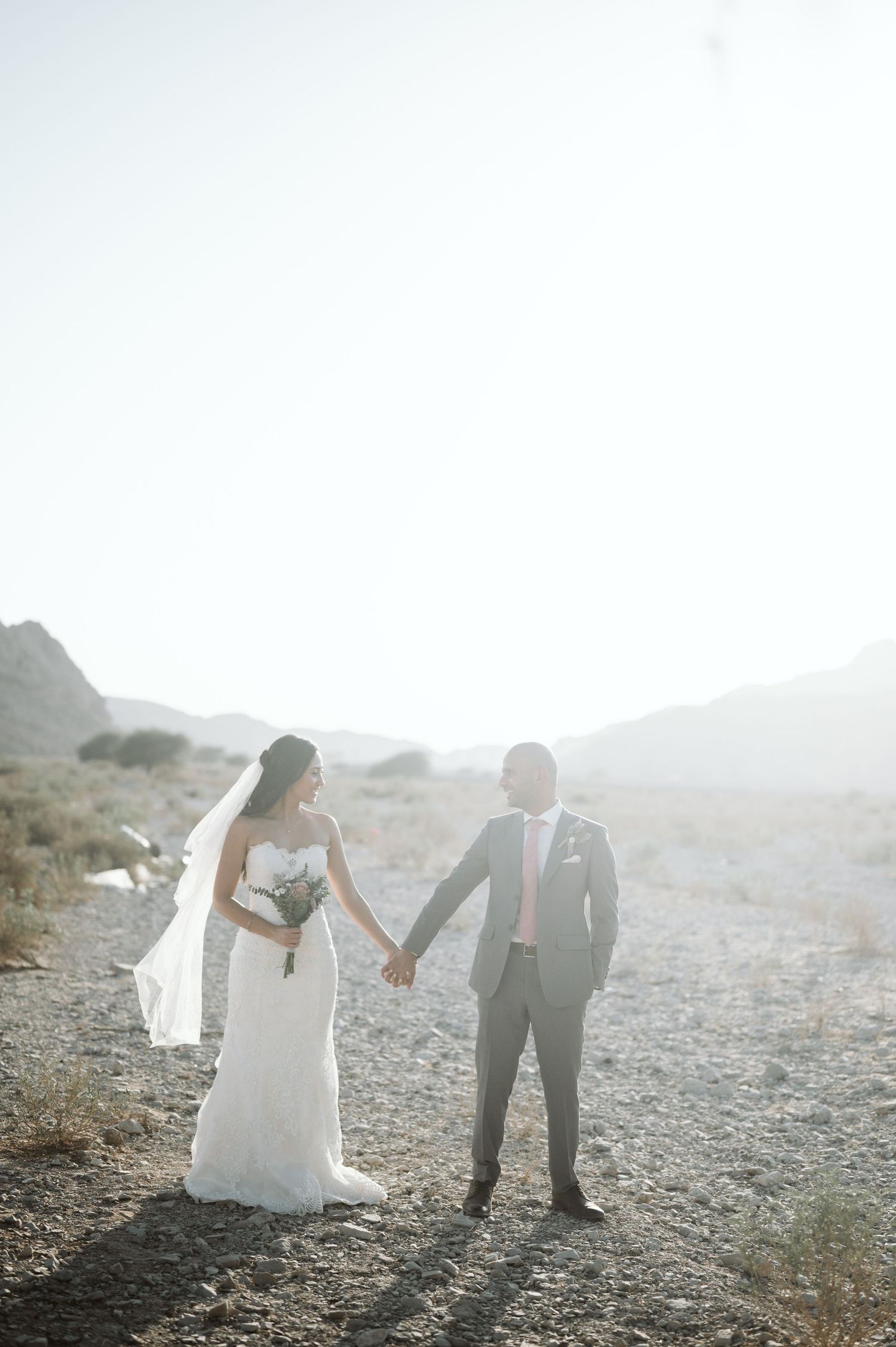 Newlyweds hold hands in desert setting with sunlight creating dramatic silhouette.