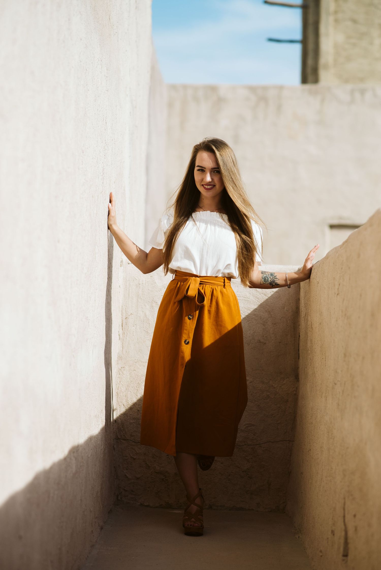 Woman in white top and orange skirt poses playfully in sunlit corridor.