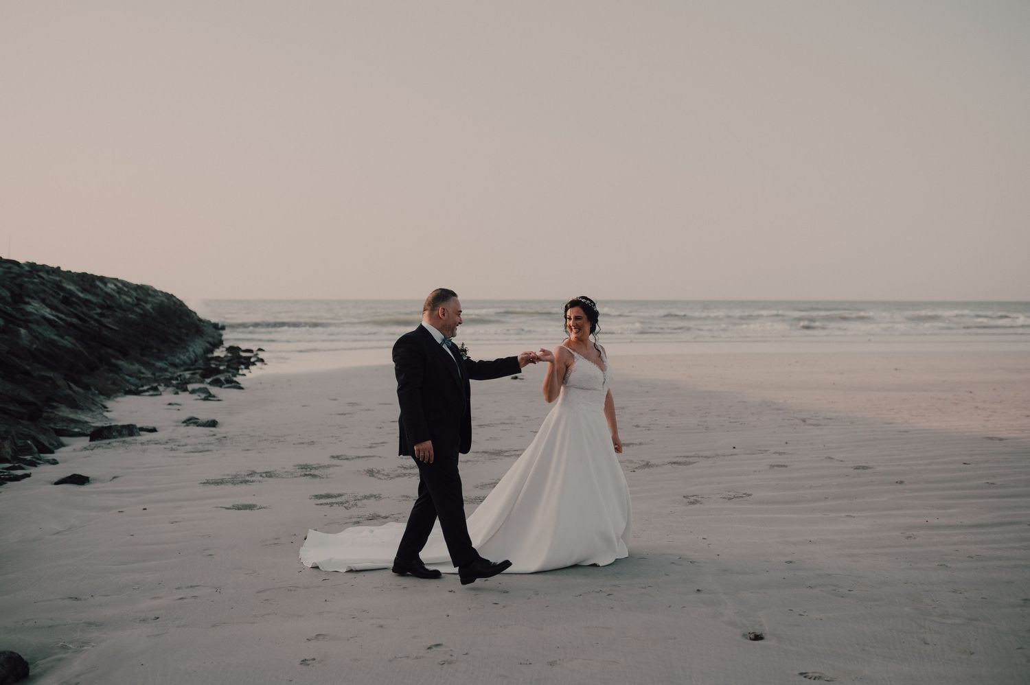 Newlyweds walk hand in hand along a serene beach at sunset.