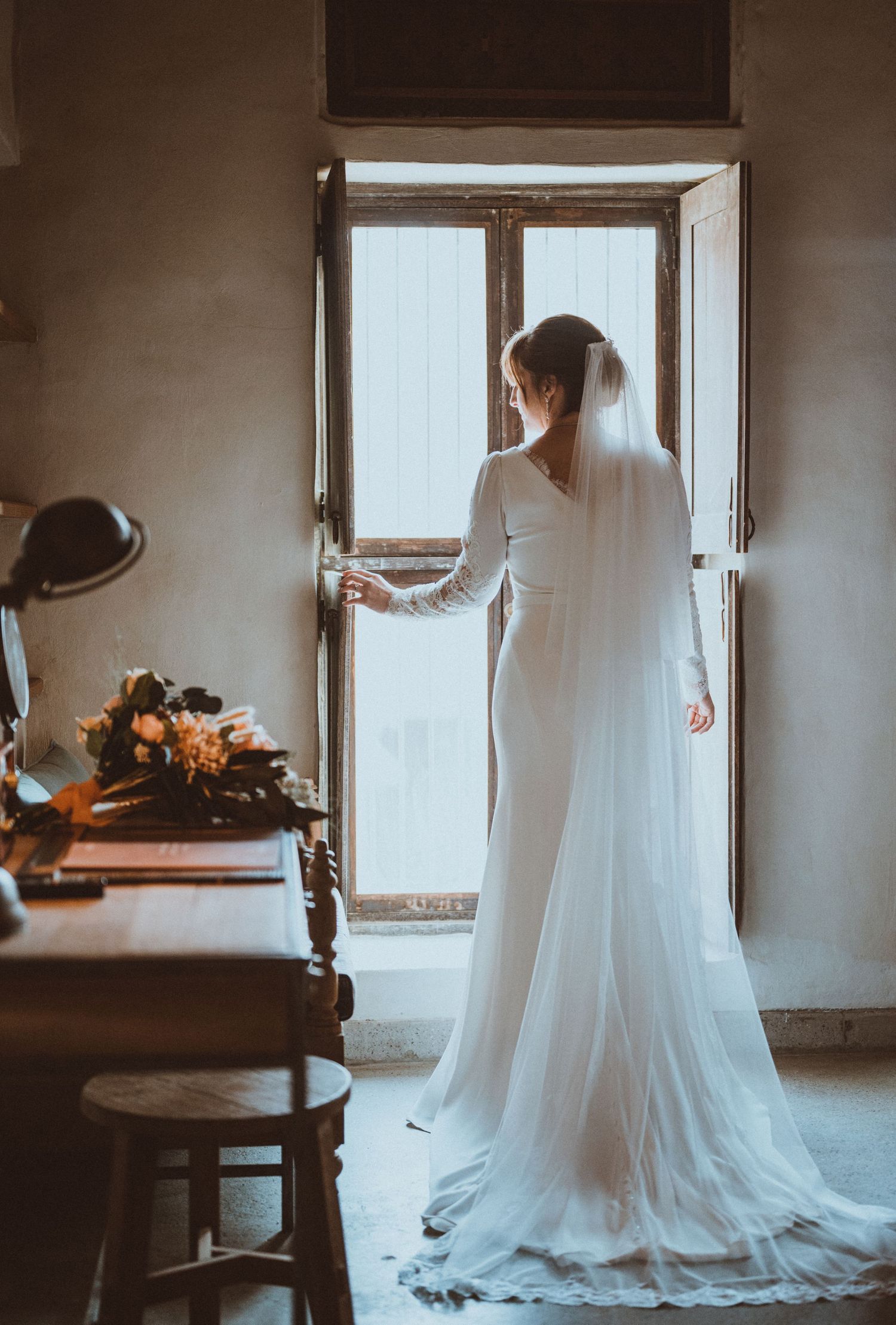 A flowing white wedding dress is photographed in soft window light against minimalist interior walls.