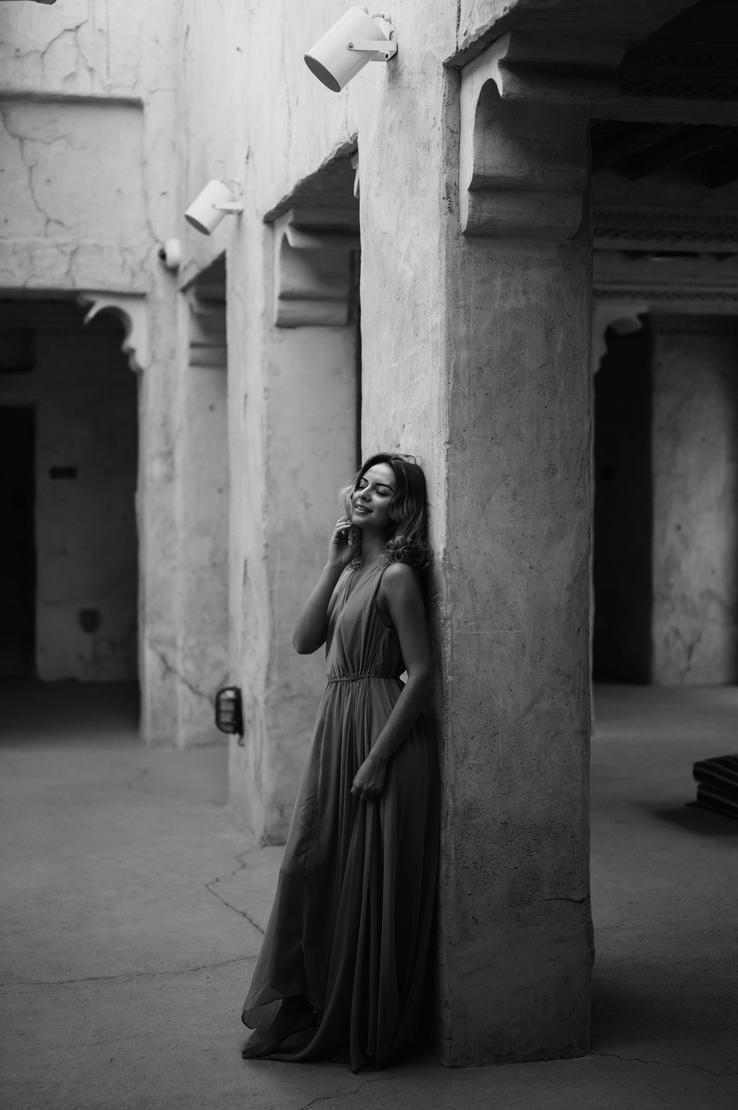 A figure in a flowing black dress poses dramatically between concrete pillars in a moody black and white photo.