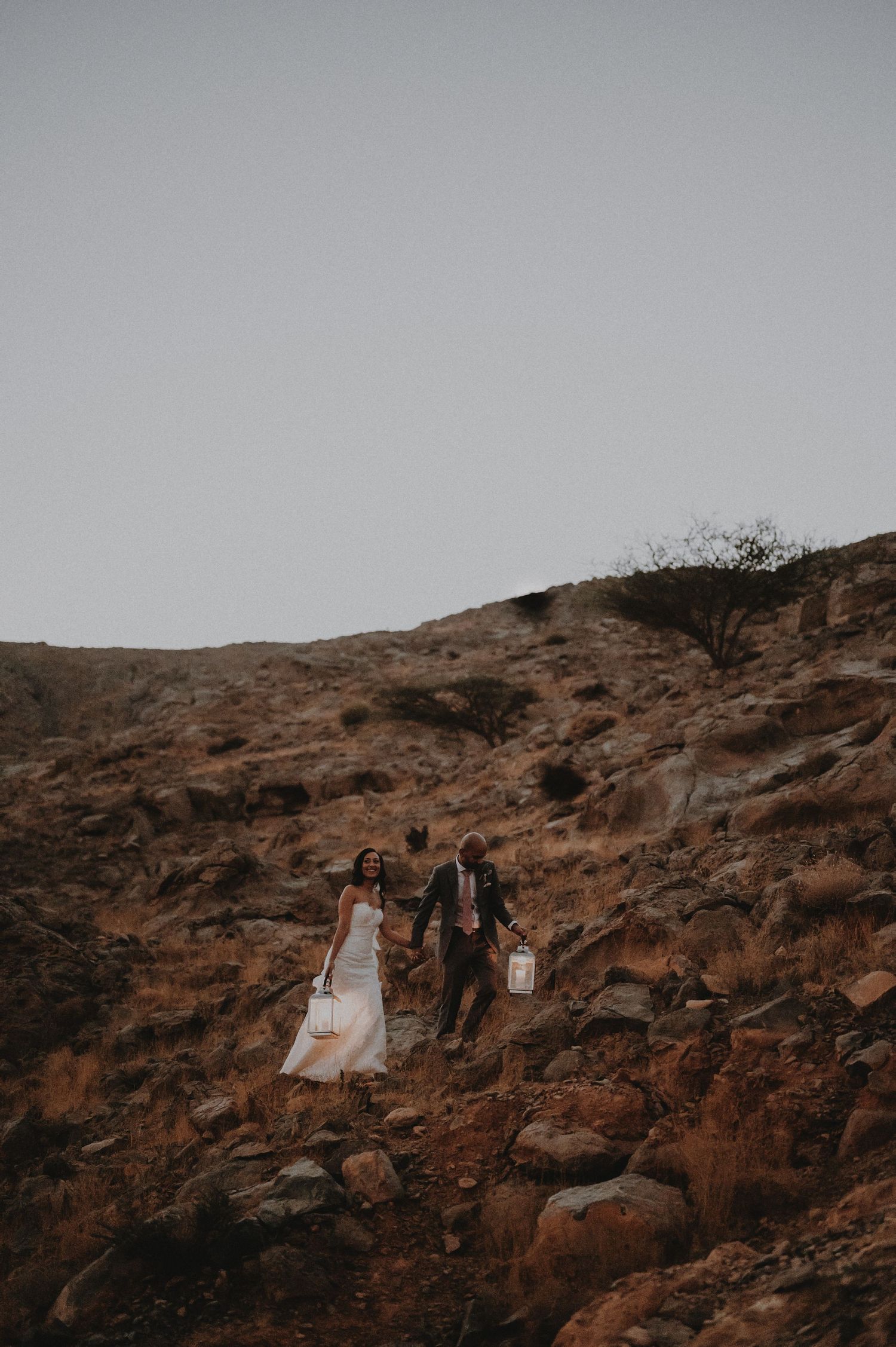 Couple sits on desert rocks at sunset holding glowing lanterns against rugged landscape.