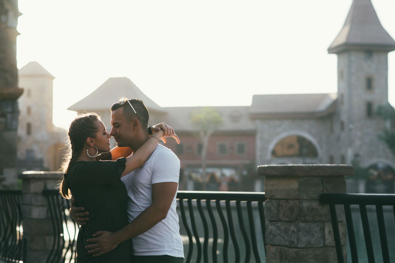 Two people share a romantic moment while standing on a bridge railing overlooking a cityscape at sunset.