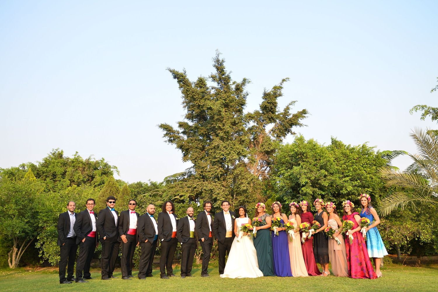 Wedding party poses outdoors in colorful formal attire on a sunny day with green trees in background.