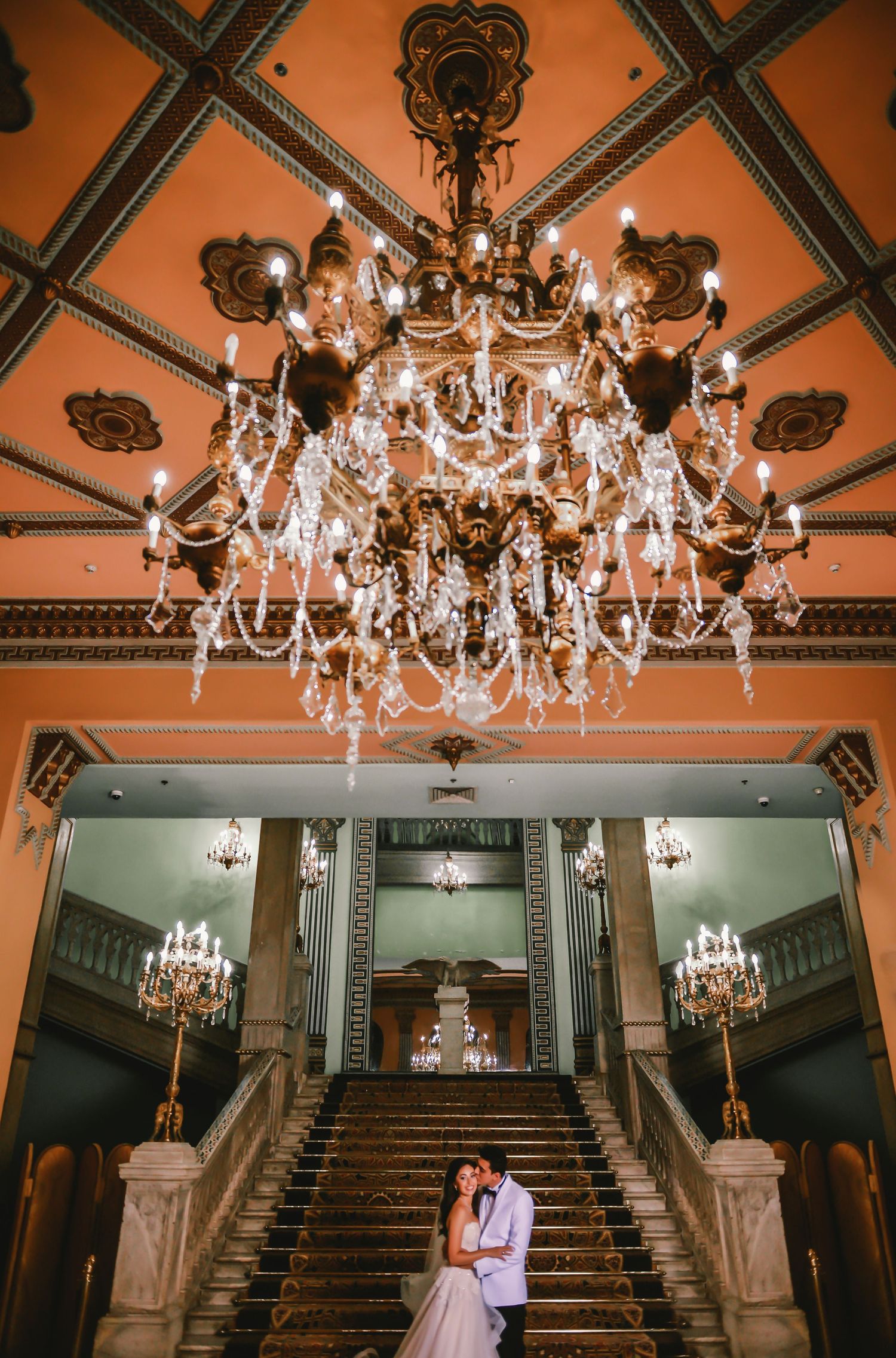 Elegant crystal chandeliers hang from an ornate ceiling above a grand staircase.