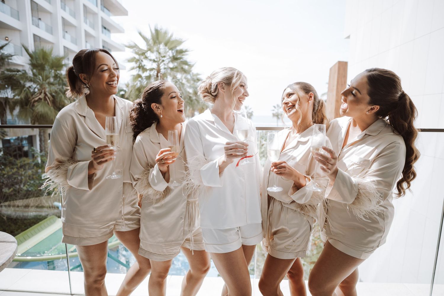 Group wearing matching white outfits laughing and celebrating together by a pool.
