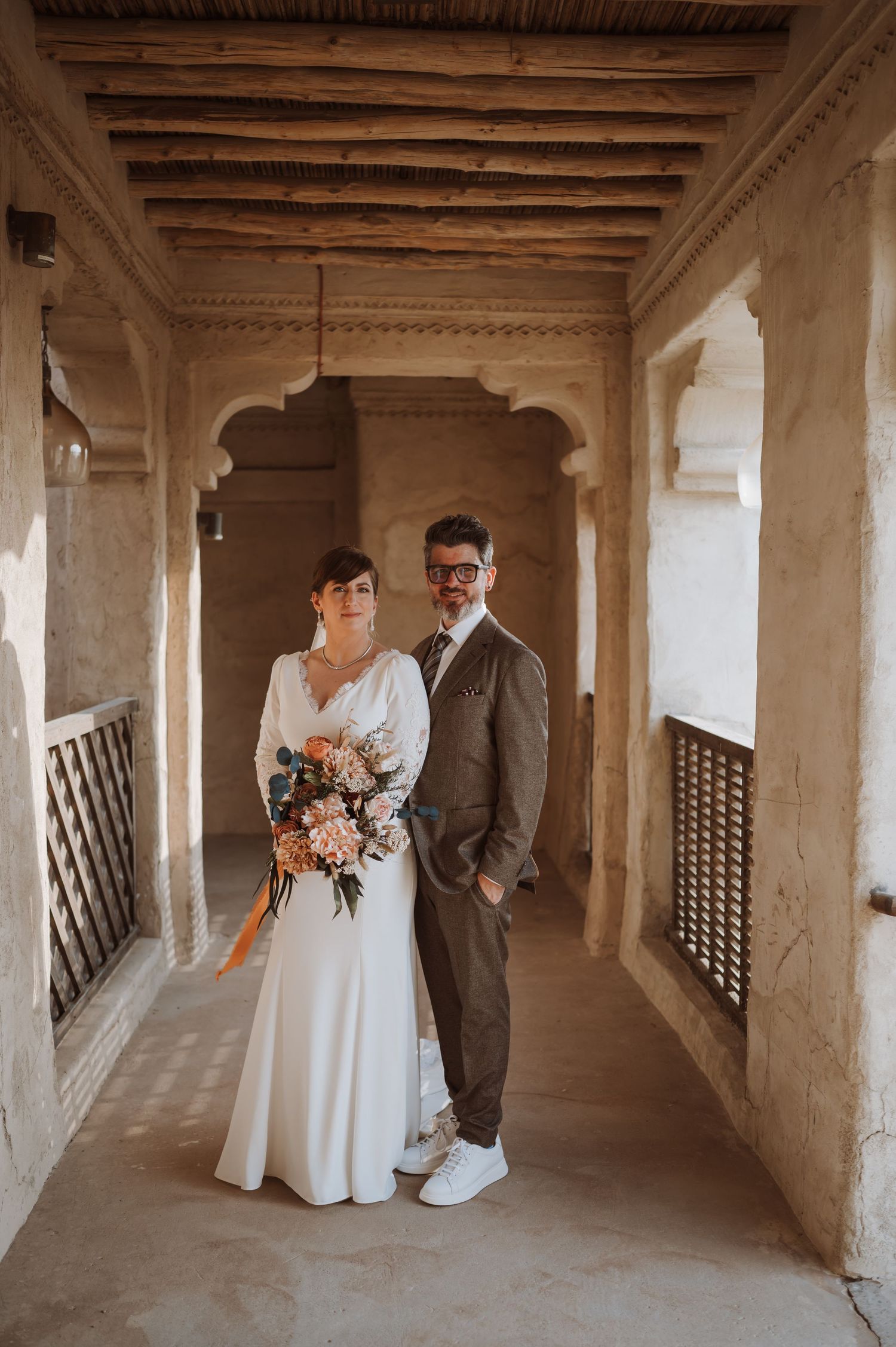 Newlyweds pose in a rustic stone corridor with white latticed windows and exposed wooden beams.