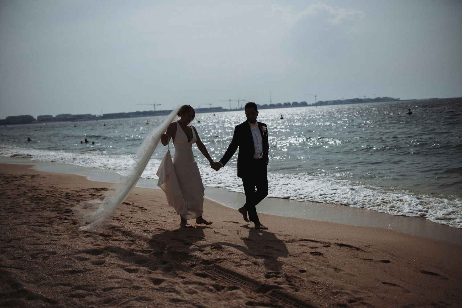 Wedding couple strolls on sandy beach with dramatic overcast sky and ocean waves.