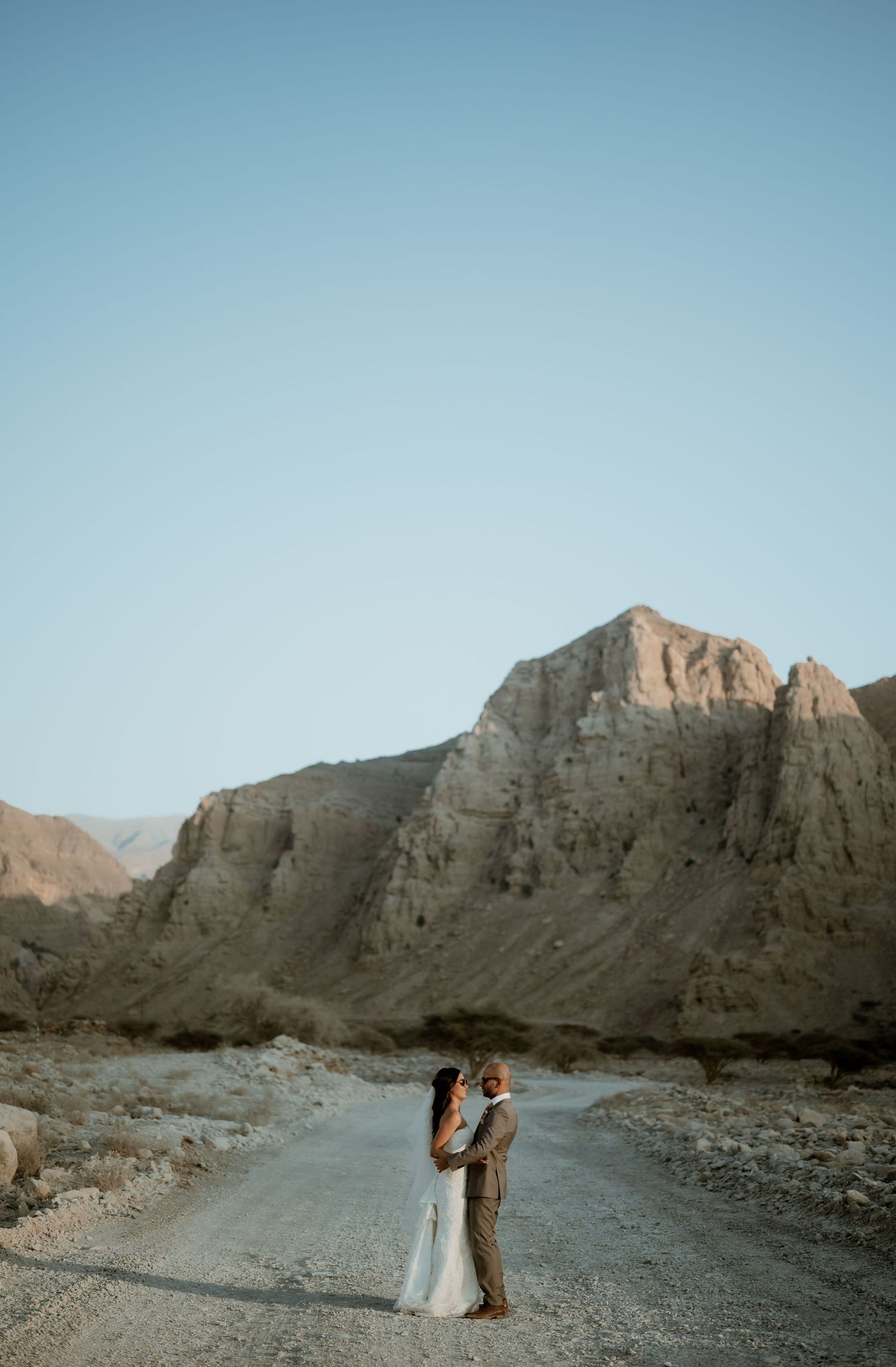 Wedding couple stands on desert road with dramatic cliff face in background.