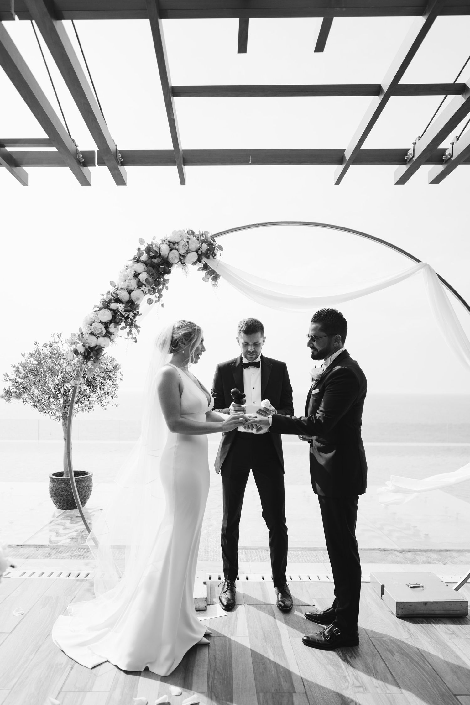 A seaside wedding ceremony unfolds under a wooden pergola with floral decorations.