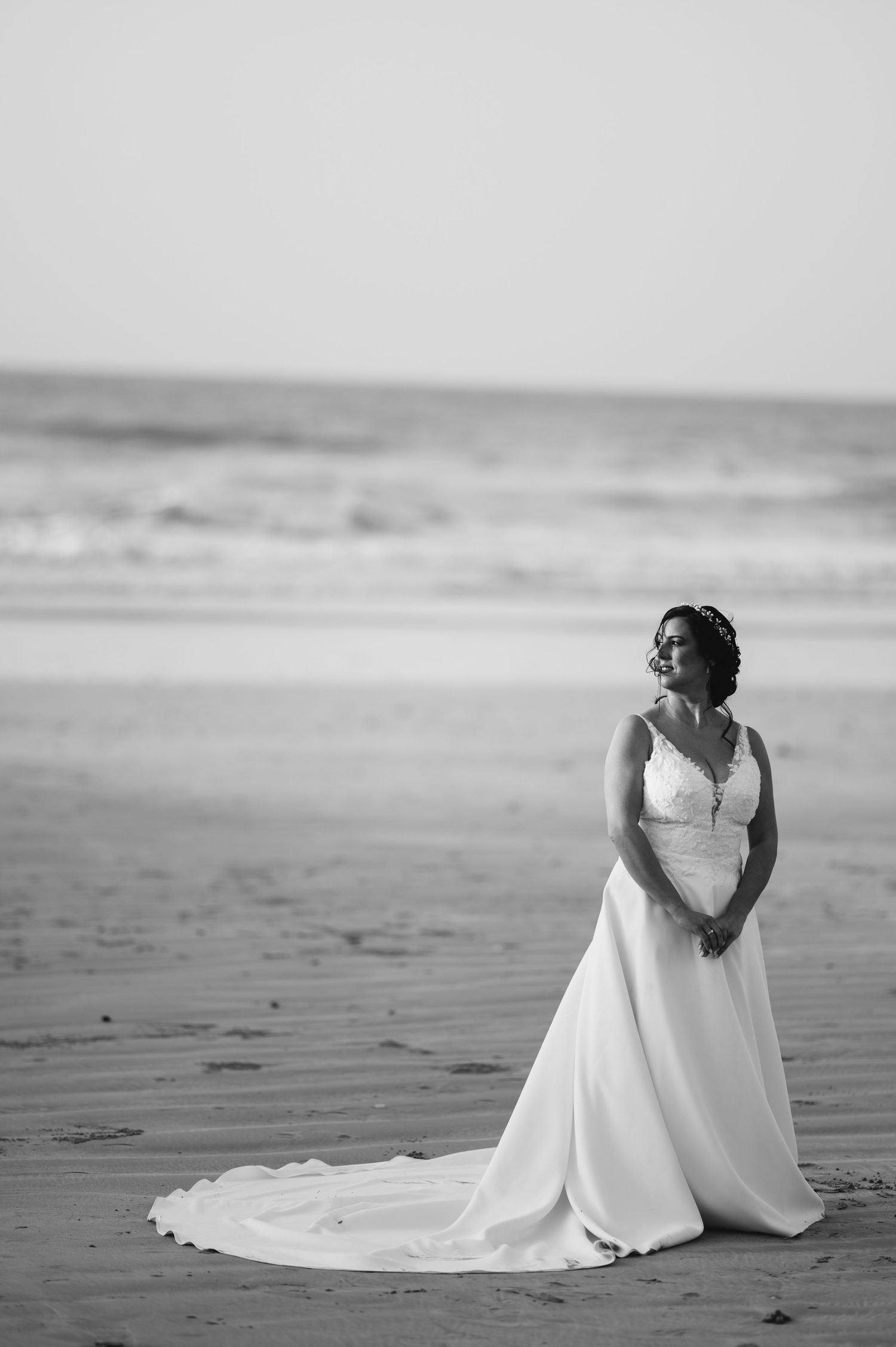 A bride in a flowing white gown poses elegantly on a sandy beach in black and white.