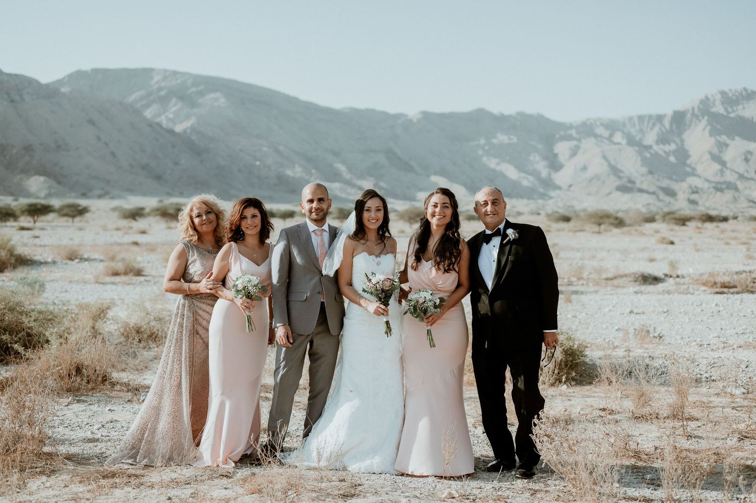 Wedding party in formal attire gathered in desert with mountain range behind them.