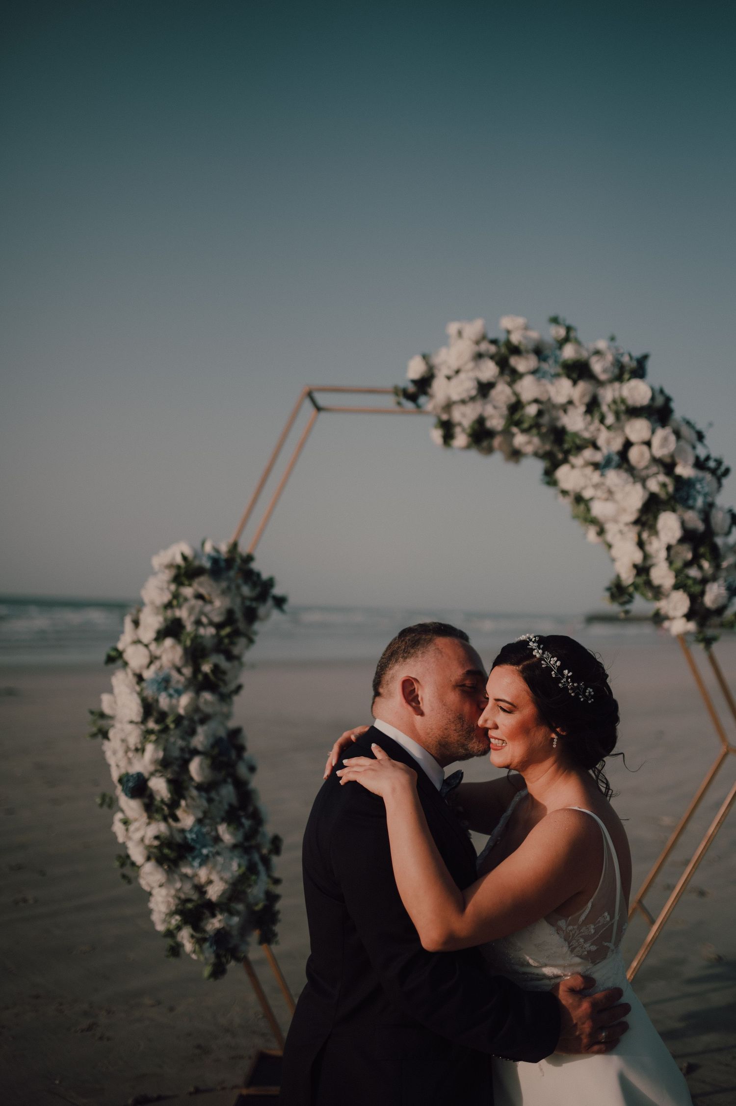 Romantic beach wedding scene with floral arch and ocean backdrop at dusk.