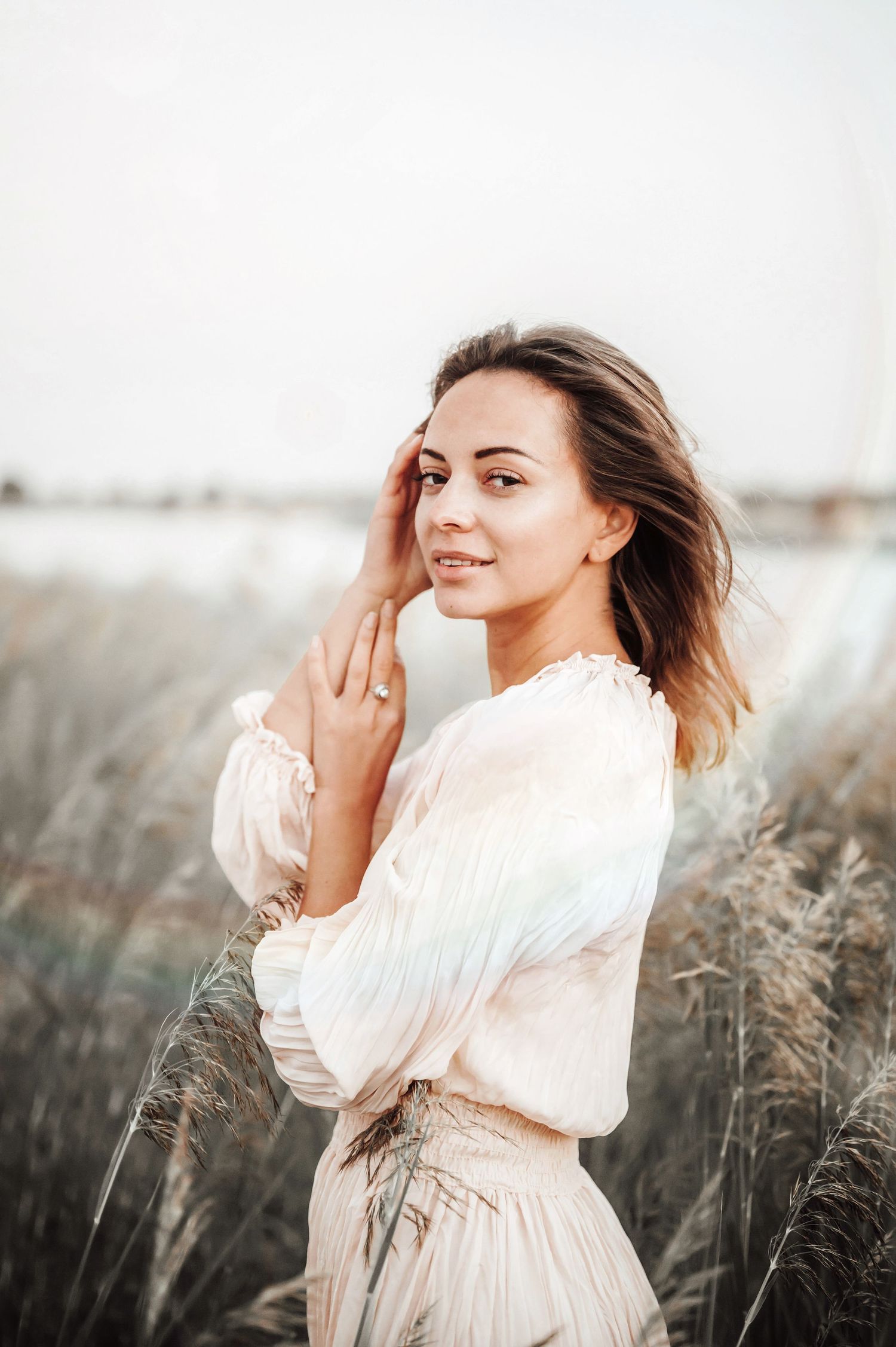 Woman in white flowing dress poses gracefully in a field of tall grass at sunset.