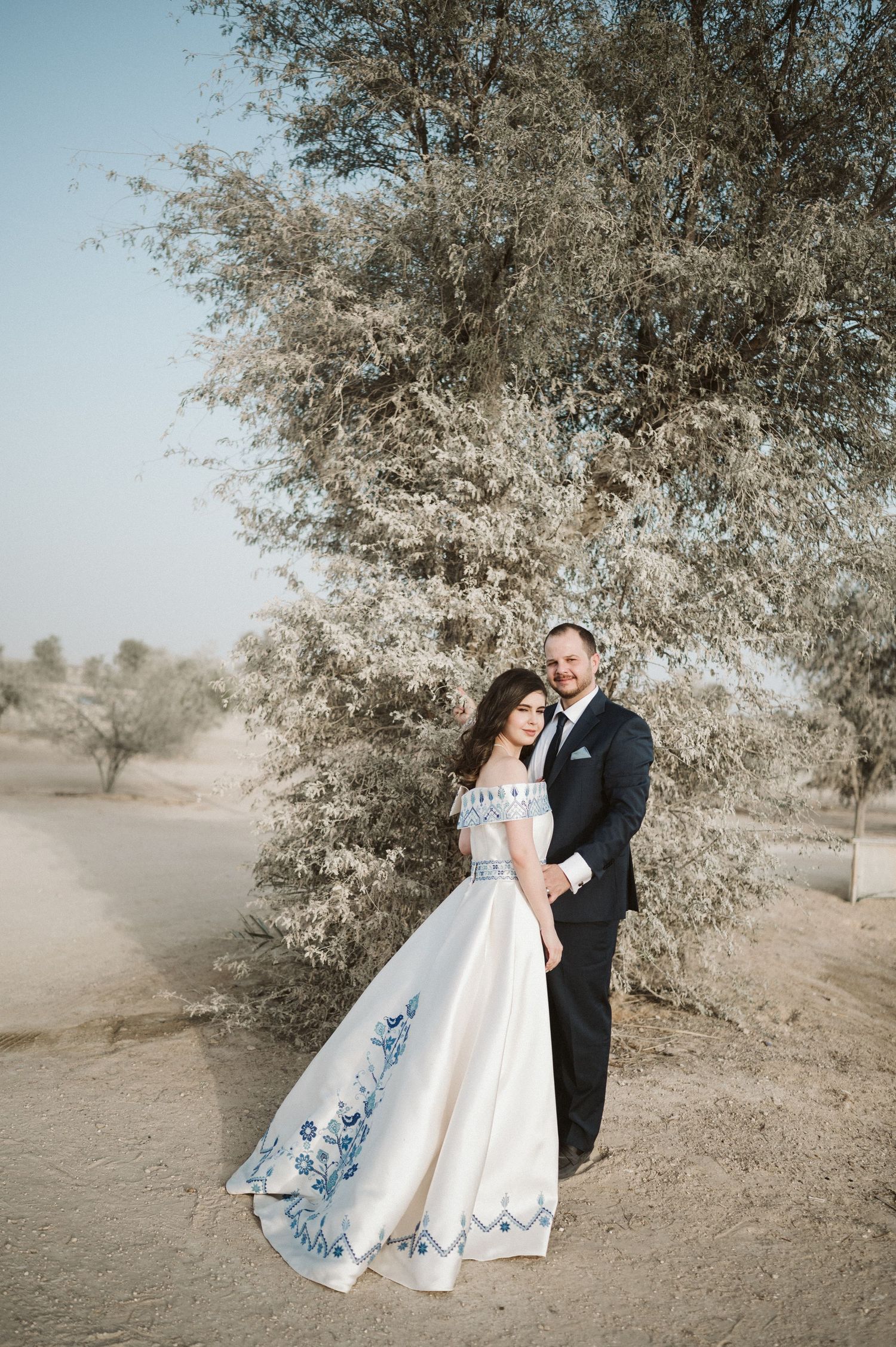 A desert wedding photoshoot features a couple posing among scattered trees in soft lighting.