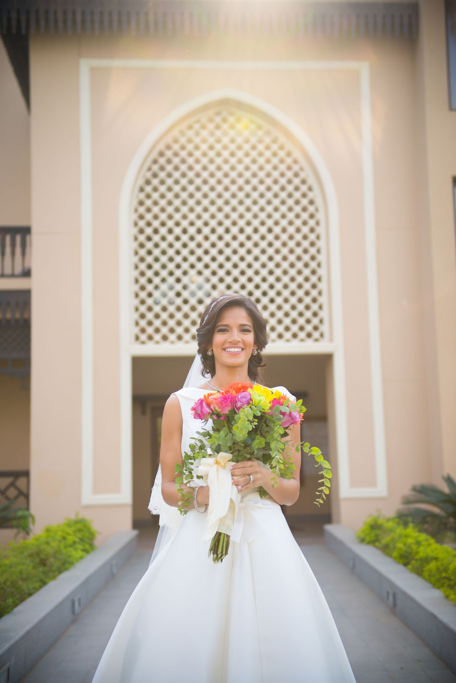 A bride in a white wedding dress holds a vibrant bouquet of pink and yellow flowers against an arched colonnade.