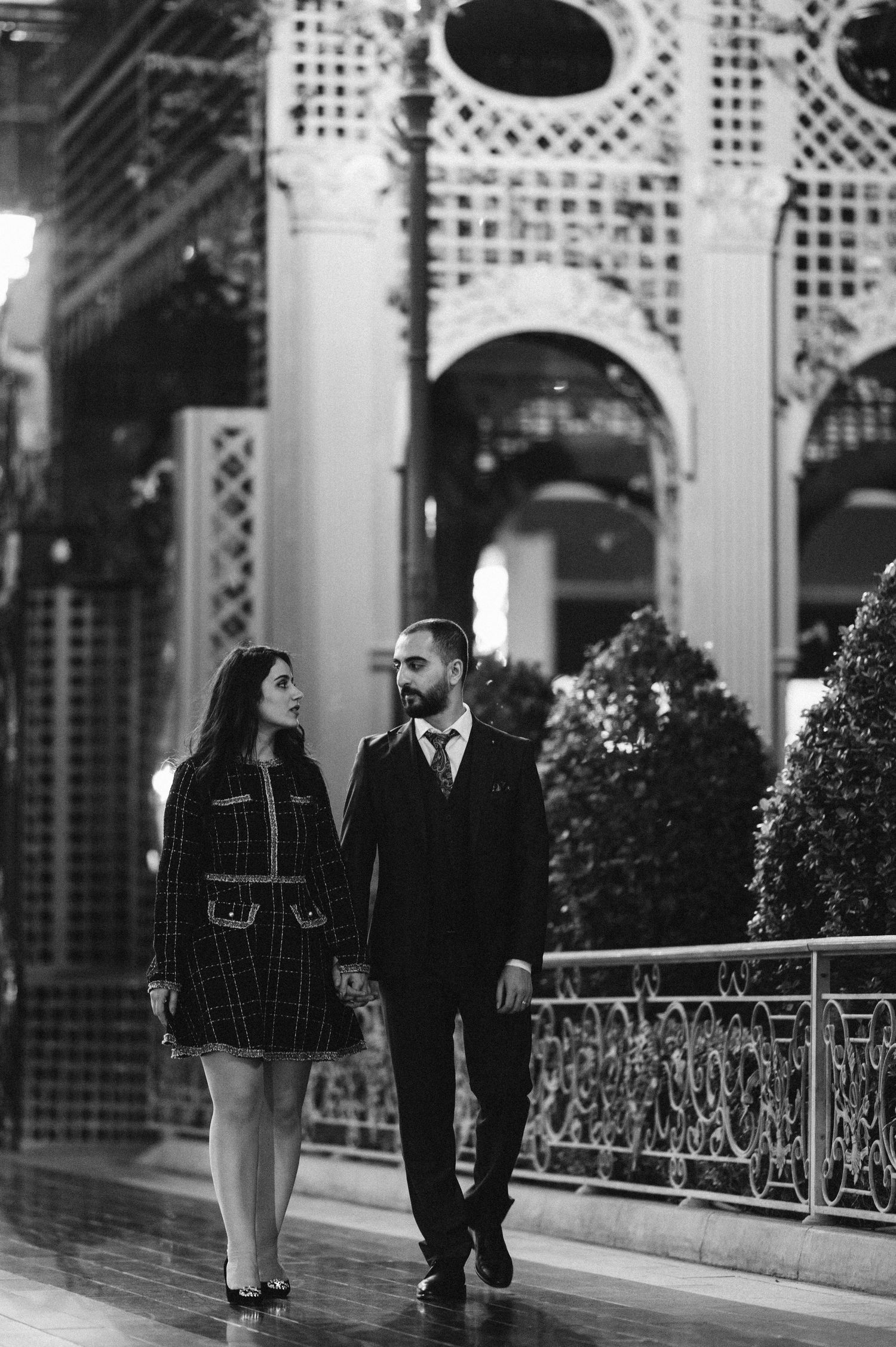 A romantic black and white photo of a couple walking together along an ornate architectural pathway.